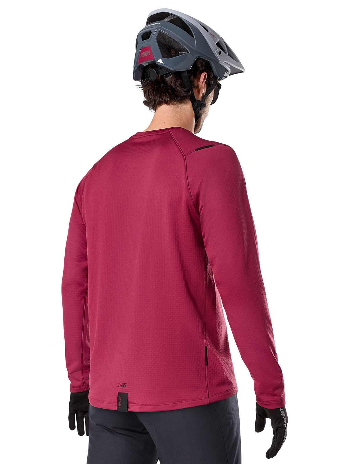 Alpinestars A-Dura Thermal Jersey, jersey de ciclismo de montaña de manga larga, granate, vista trasera en modelo con casco de MTB gris y guantes negros, con cola extendida y pequeños detalles de logotipo, diseñado para ciclismo de montaña en climas fríos