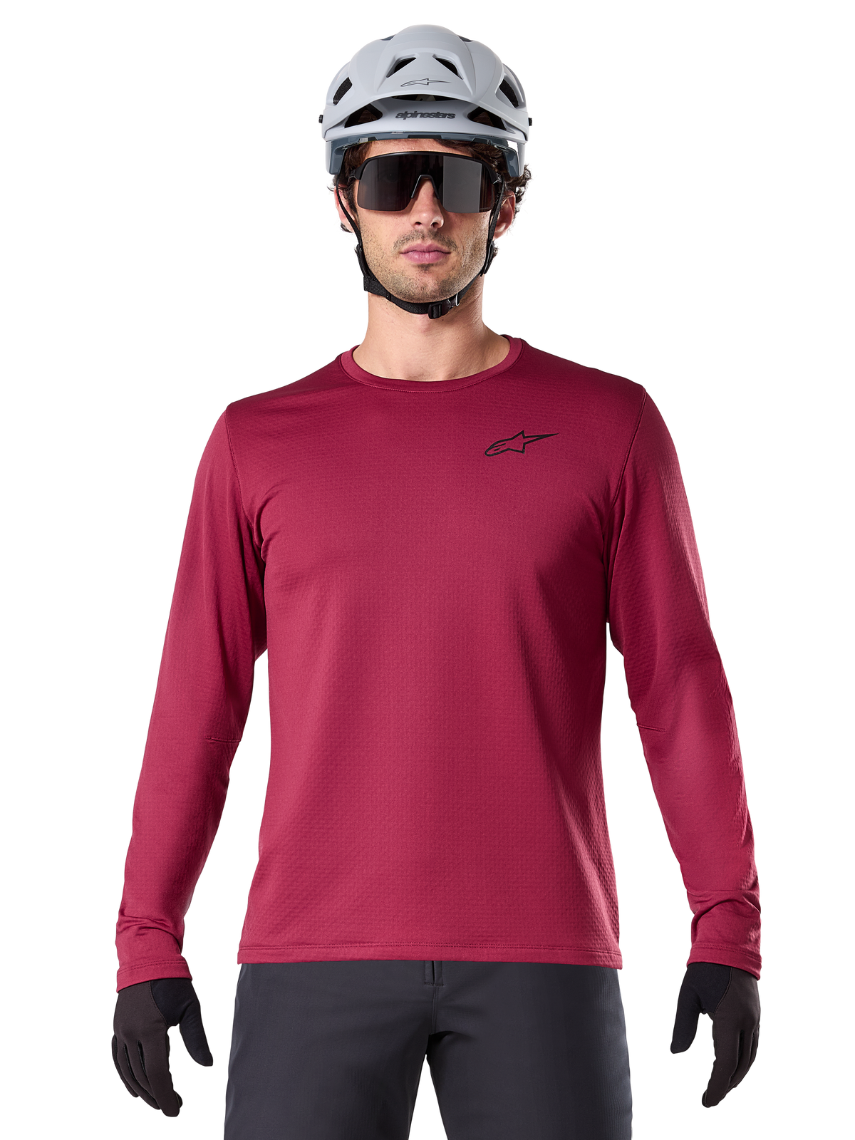 Ciclista masculino con el Jersey Térmico A-Dura de Alpinestars, jersey de MTB de manga larga, color granate, combinado con un casco gris y gafas de sol negras, presenta un pequeño logotipo de estrella negro en el pecho y tejido de vellón aislante para ciclismo de montaña en climas fríos