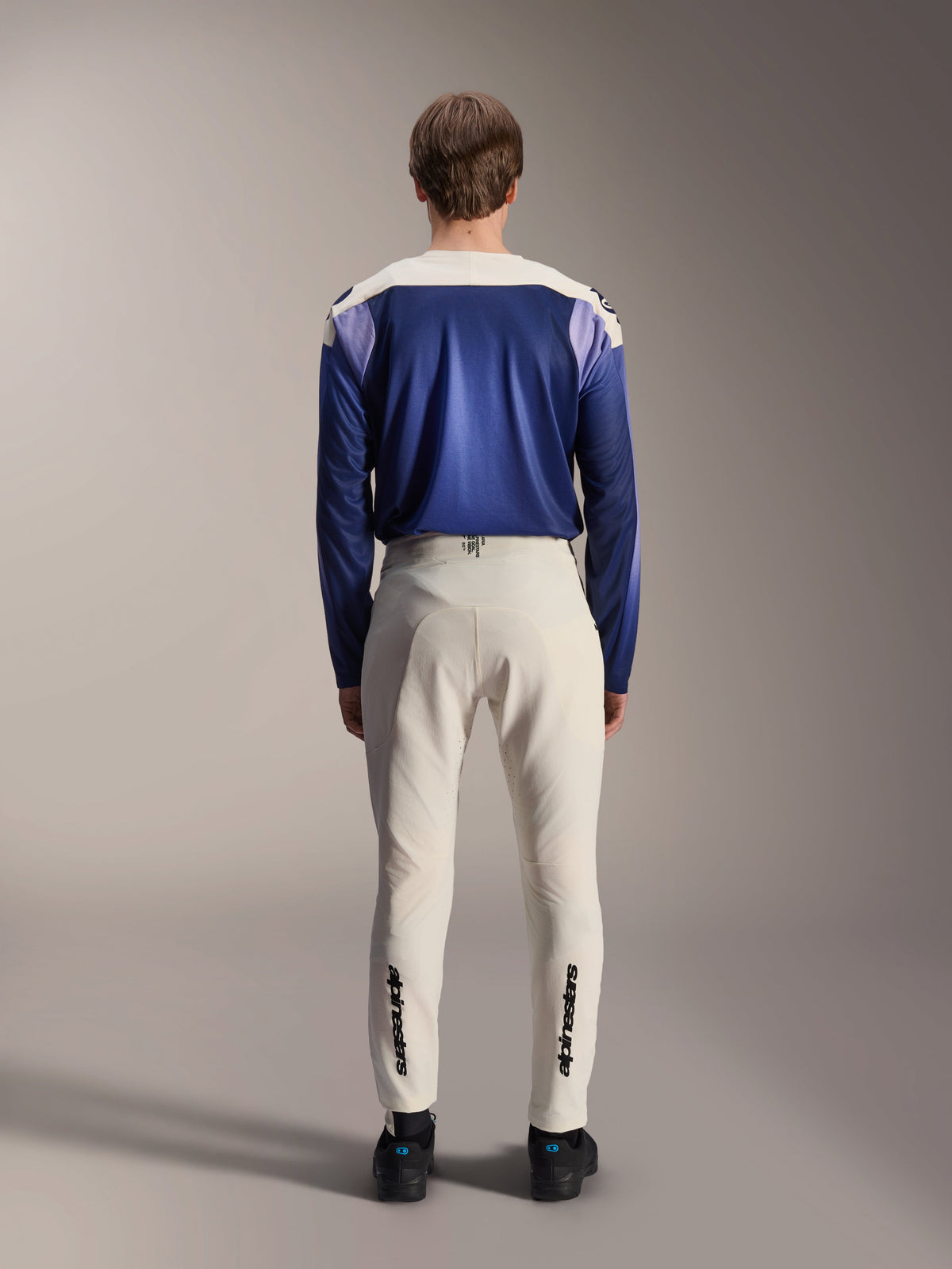 Alpinestars A-Aria Polartec Maker Elite Long Sleeve Jersey, Baja Blue and off-white, back view of model wearing technical mountain bike gear including off-white pants with Alpinestars branding on the lower legs, performance cycling outfit for off-road trails