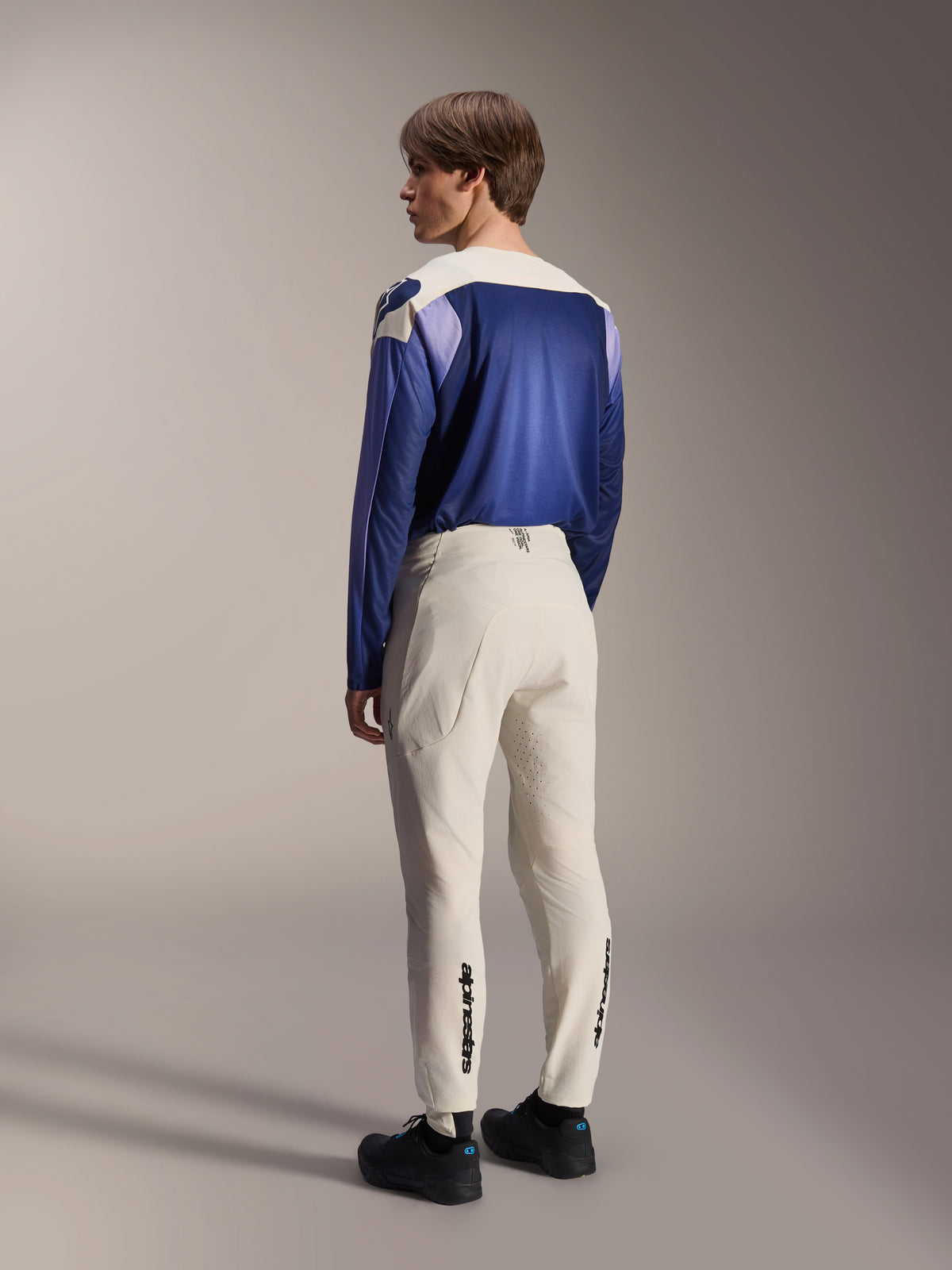 Alpinestars A-Aria Elite Pants, MTB Pants, Ecru off-white, rear view worn by a model, featuring black wordmark branding on the lower legs and laser perforations for ventilation, paired with a blue and purple long-sleeve jersey and black shoes, professional mountain bike gear