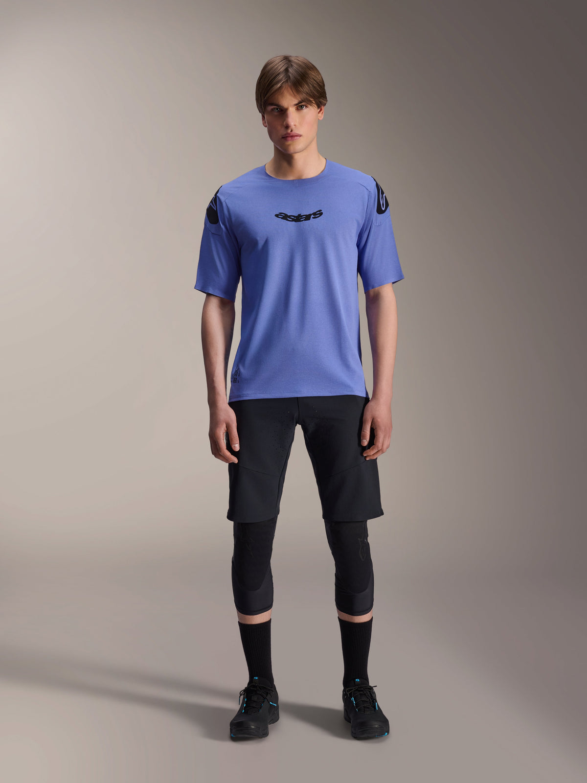 Modelo masculino con el jersey de manga corta Alpinestars A-Aria Polartec Elite en color Baja Blue, logotipo astars negro en el pecho, combinado con pantalones cortos de MTB negros, rodilleras y zapatillas de ciclismo negras, toma de estudio de ropa para bicicleta de montaña