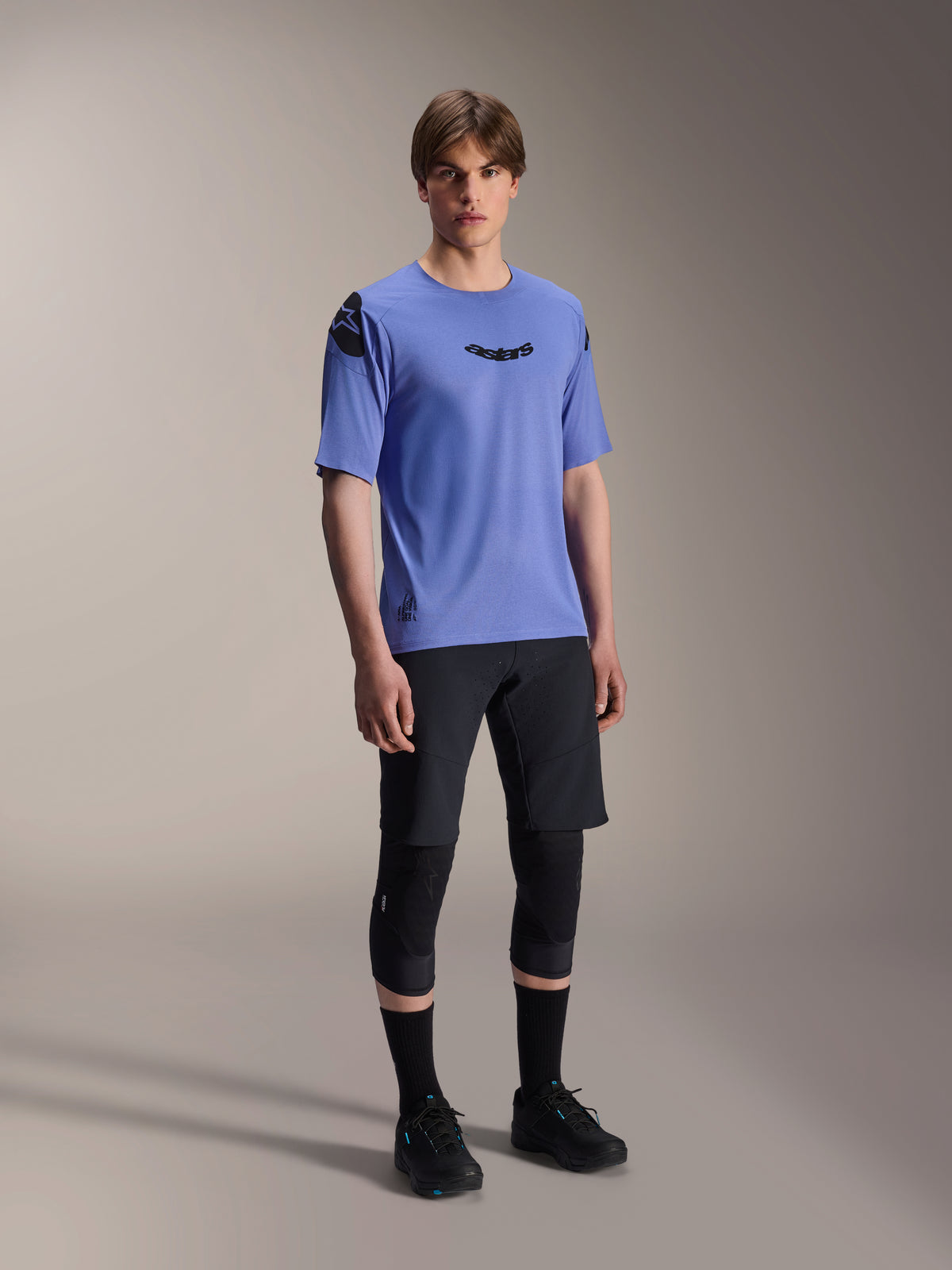 Modelo masculino vistiendo shorts Alpinestars A-Aria Elite en color Black Titanium y jersey A-Aria Polartec Elite SS en púrpura claro, combinados con rodilleras y zapatillas de MTB negras; equipamiento de ciclismo profesional para rendimiento off-road.