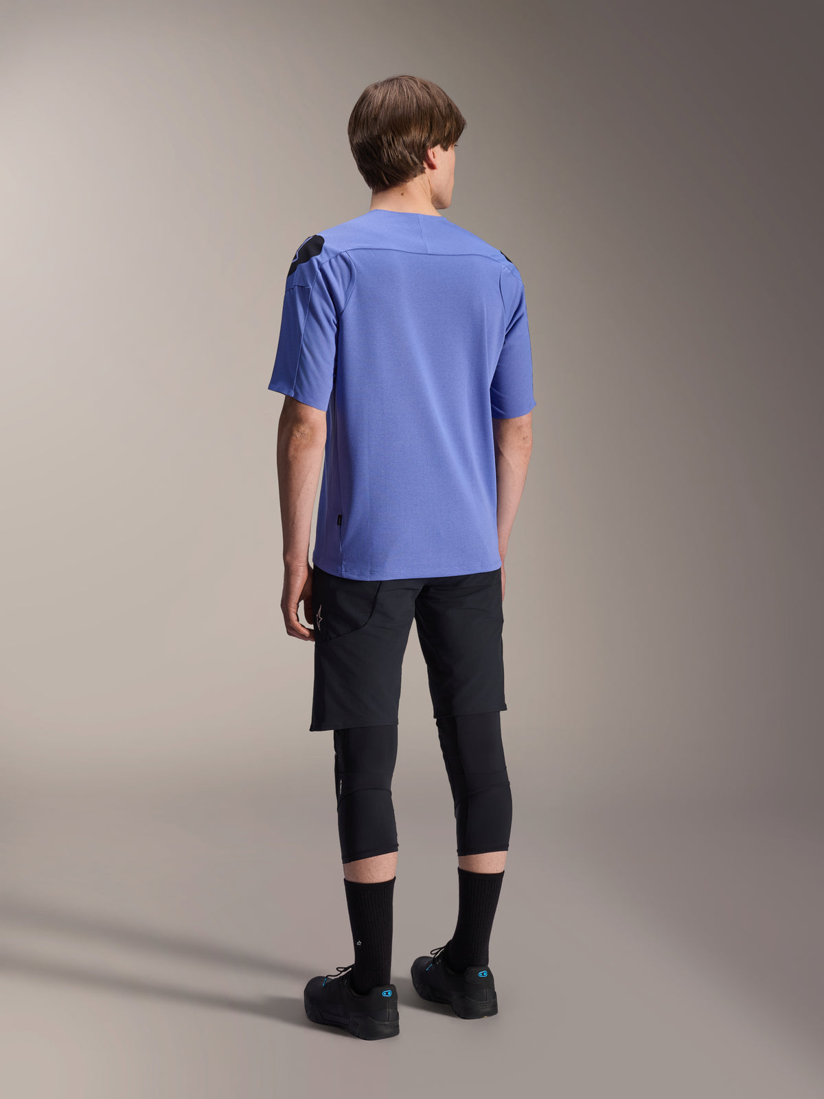 Alpinestars A-Aria Lite Short, shorts de ciclismo de montaña negros, combinados con un jersey de manga corta azul A-Aria Polartec Elite y rodilleras negras, vista posterior de un ciclista con equipamiento de ciclismo de alto rendimiento para senderos todoterreno.