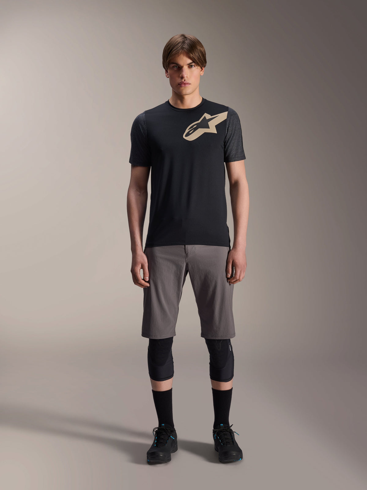 Jersey de manga corta Alpinestars A-Dura Lipan, negro con mangas gris jaspeado y logotipo Astars color canela en el pecho, usado por un modelo masculino con pantalones cortos de ciclismo de montaña grises y rodilleras negras, equipamiento técnico para MTB.