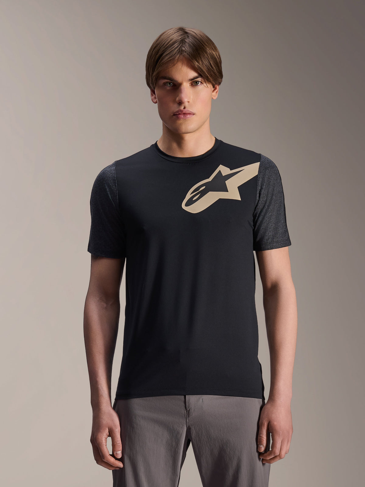 Modelo masculino con jersey de manga corta Alpinestars A-Dura Lipan, jersey de MTB negro con logo de estrella beige y mangas gris oscuro, combinado con pantalones de ciclismo grises, indumentaria de rendimiento casual