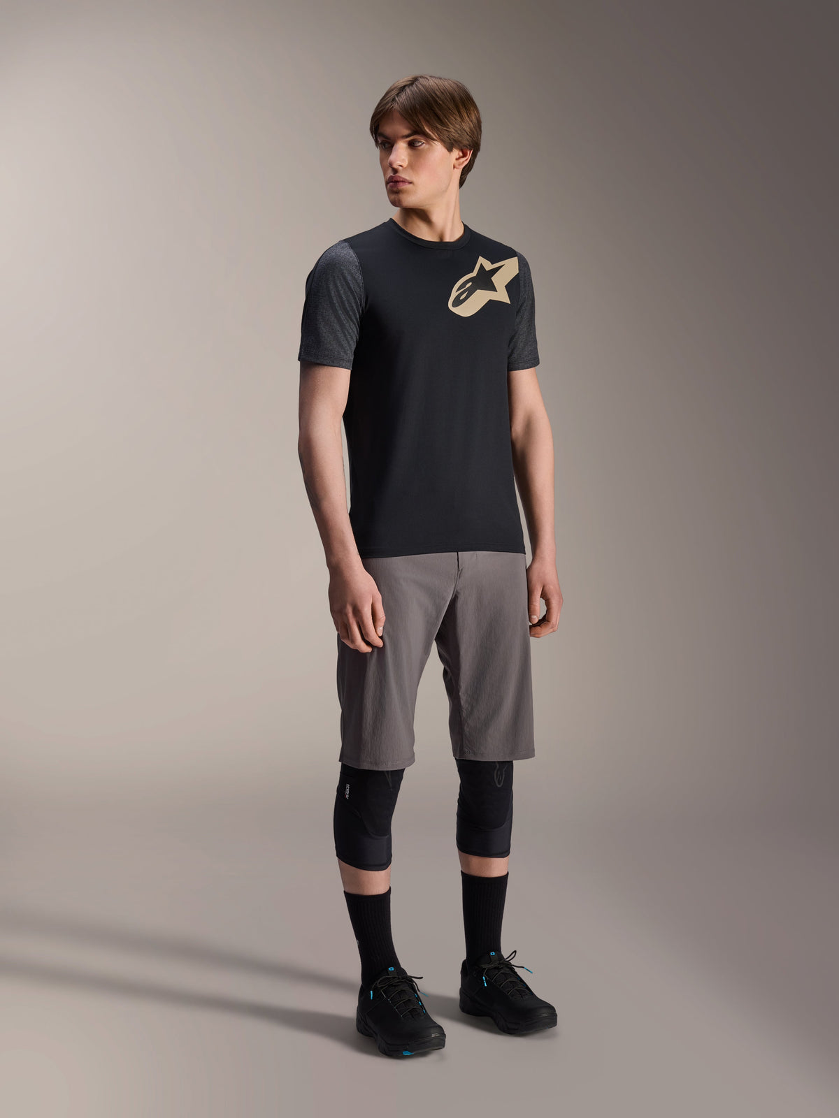 Modelo masculino luciendo el Jersey de manga corta Alpinestars A-Dura Lipan en color negro, con mangas gris oscuro y logotipo de Astars en color canela en el pecho, combinado con pantalones cortos grises de ciclismo de montaña, rodilleras negras y zapatillas de ciclismo negras, equipamiento coordinado de MTB