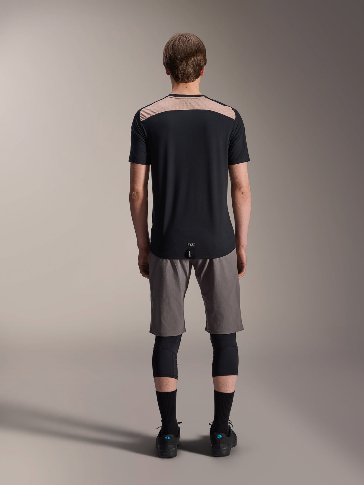 Alpinestars A-Dura Lipan Jersey - Manga corta, negro con panel superior de la espalda en color tostado, vista trasera que muestra la cola más larga, usado con pantalones cortos de MTB grises y rodilleras negras, ropa de alto rendimiento para bicicleta de montaña