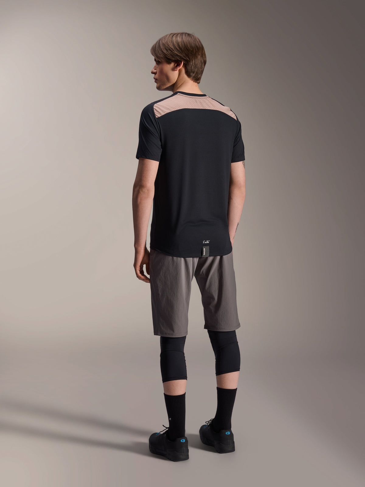 Camiseta de manga corta Alpinestars A-Dura Lipan en negro, vista trasera en modelo que muestra el panel de malla perforada marrón claro en la parte superior de la espalda, combinada con pantalones cortos de ciclismo de montaña grises y rodilleras negras para un conjunto completo de trail.