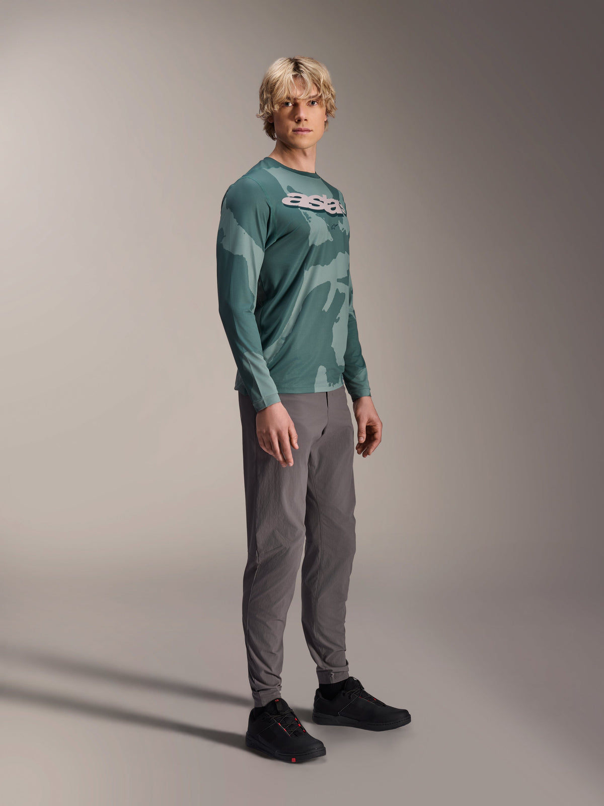 Alpinestars A-Dura Hollow Camo Long Sleeve Jersey, Silver Pine Camo, worn by a model with grey pants and black shoes, featuring a green camouflage pattern and a bold white ASTARS logo on the chest, high-performance MTB apparel