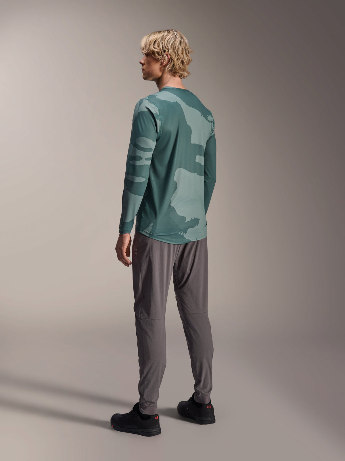 Alpinestars A-Dura Hollow Camo Long Sleeve Jersey, Silver Pine Camo, back view of model wearing green camo jersey with grey pants and black shoes, showing drop tail and camo pattern, performance mountain bike gear
