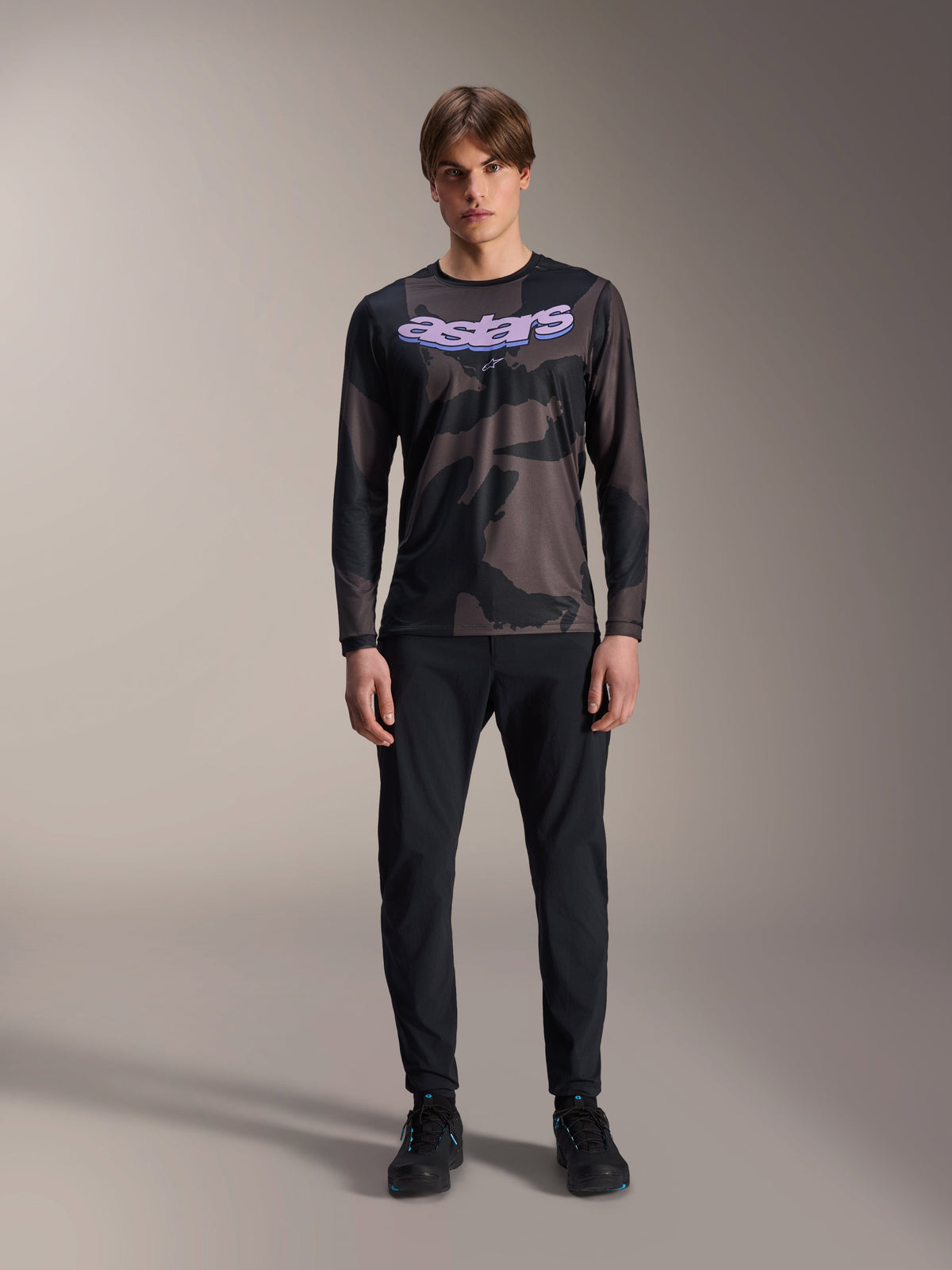 Model wearing Alpinestars A-Dura Hollow Camo Long Sleeve Jersey in Black Camo, featuring a purple 'astars' logo on the chest, paired with black pants and black cycling shoes, mountain bike riding gear