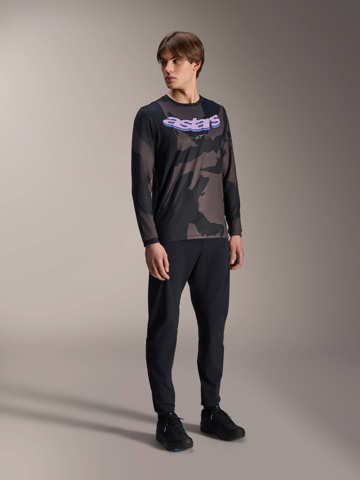 Male model wearing Alpinestars A-Dura Hollow Camo Long Sleeve Jersey, Black Camo pattern with purple 'astars' chest graphic, paired with black technical pants and black shoes, casual MTB gear
