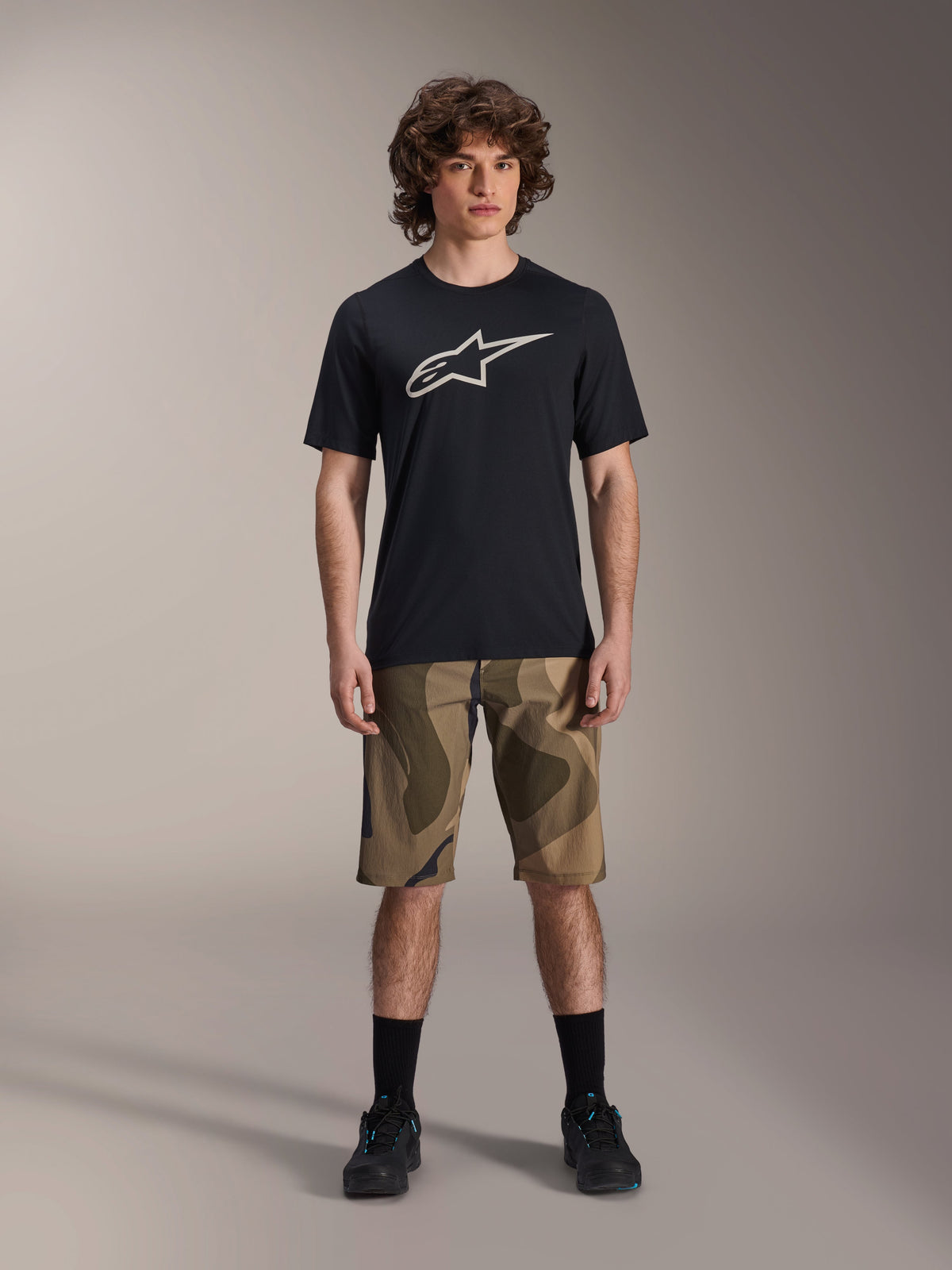 Jersey de manga corta Alpinestars A-Dura Astar, negro con un gran logotipo de Astars blanco en el pecho, usado por un modelo con pantalones cortos de MTB de camuflaje y zapatillas de ciclismo negras, ropa casual de mountain bike.