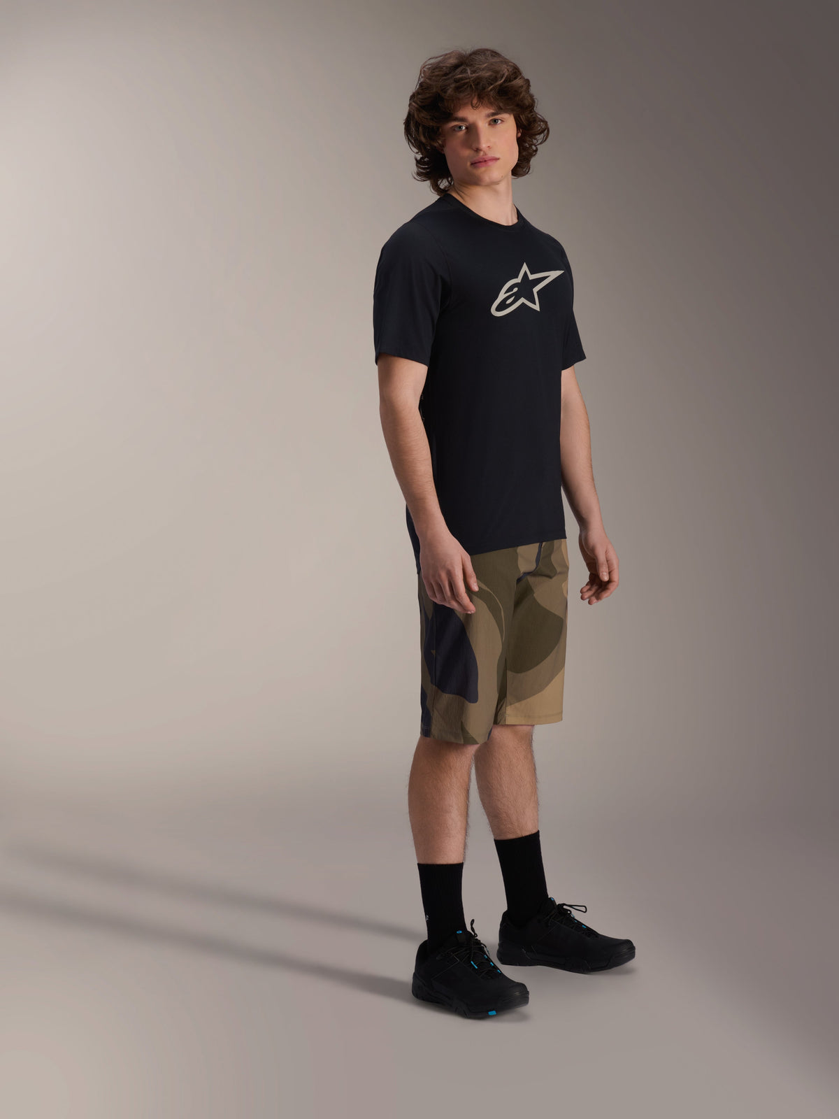 Jersey de manga corta Alpinestars A-Dura Astar, negro con logotipo blanco en el pecho, usado con pantalones cortos de camuflaje y zapatillas de MTB negras, equipamiento casual para ciclismo de montaña