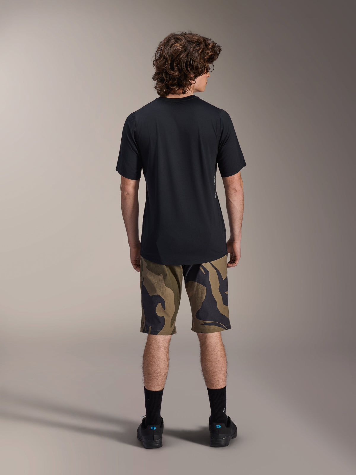 Jersey de manga corta Alpinestars A-Dura Astar, negro, vista trasera en un modelo combinado con pantalones cortos de MTB con estampado de camuflaje y calzado negro, ropa técnica casual para bicicleta de montaña