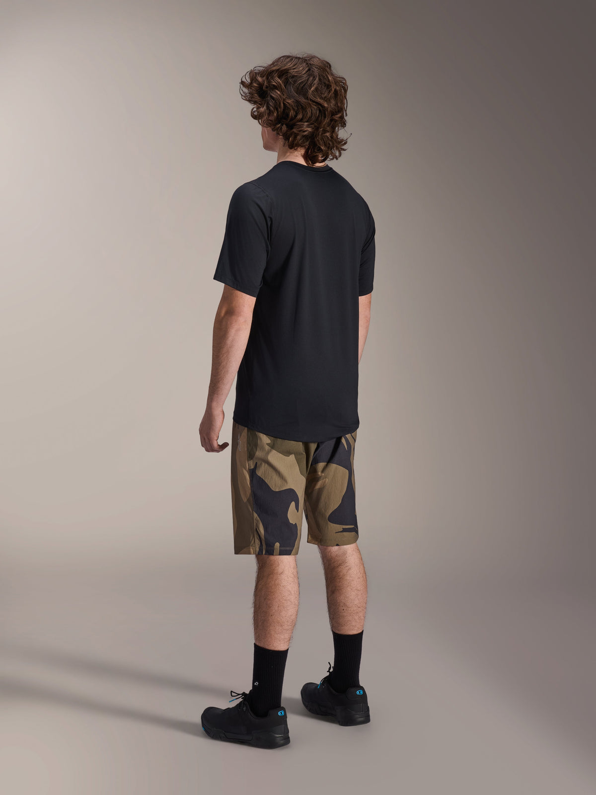 Alpinestars A-Dura Astar Jersey de manga corta en negro, visto desde atrás, combinado con pantalones cortos con estampado de camuflaje, calcetines y zapatillas deportivas negras, equipamiento casual para MTB