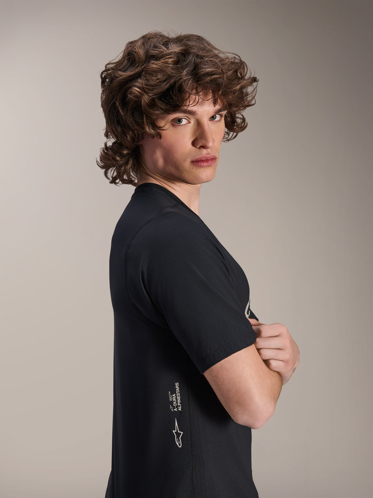Alpinestars A-Dura Astar Jersey, jersey de MTB de manga corta, negro, modelo masculino posando de perfil con los brazos cruzados, con la marca Alpinestars en blanco en el lateral inferior, diseño deportivo casual para ciclismo de montaña