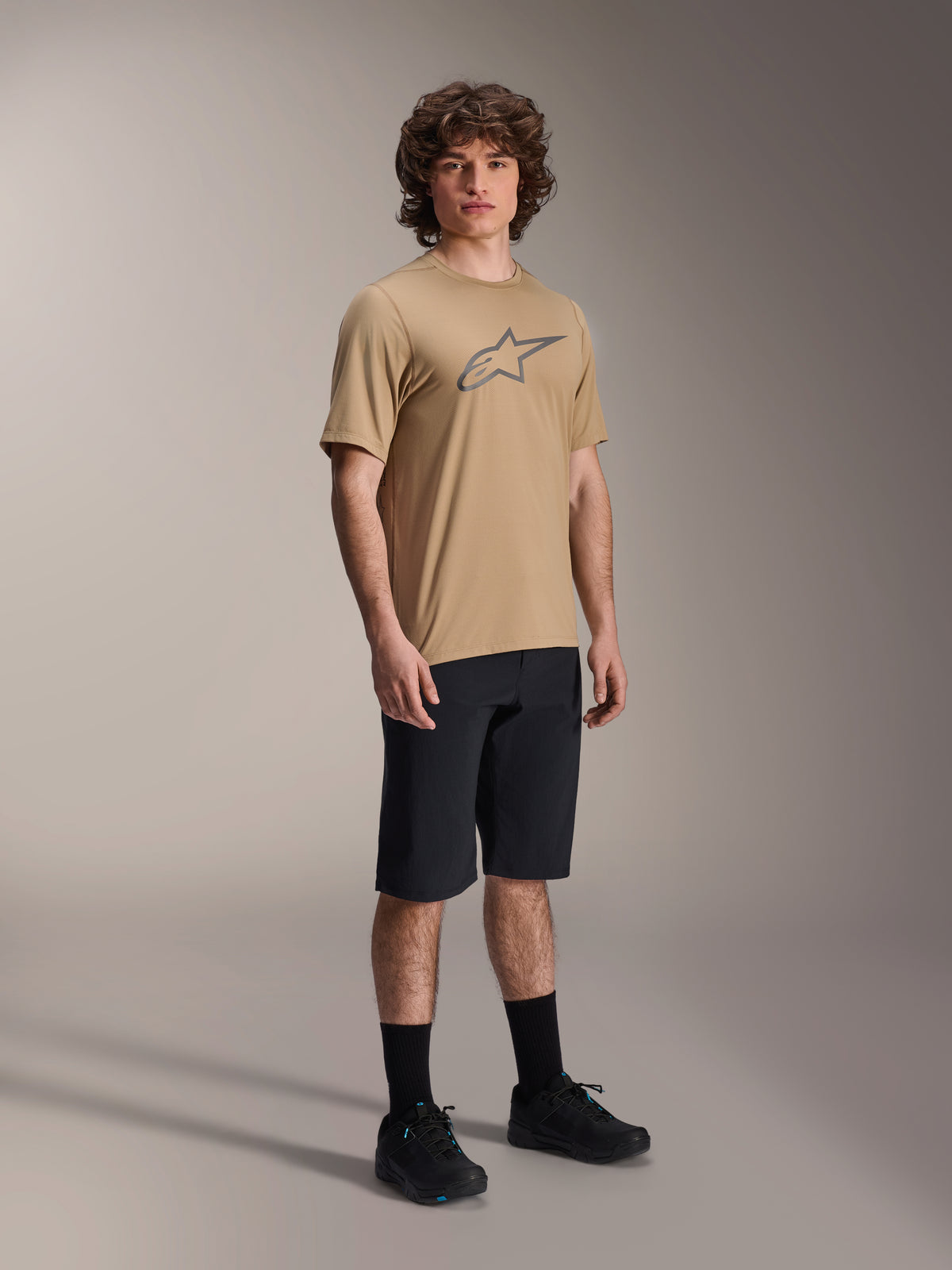Modelo masculino con shorts A-Dura Liner de Alpinestars en color negro, combinados con un jersey A-Dura SS en color tostado con un logotipo Astars gris, calcetines de ciclismo negros y zapatillas de bicicleta de montaña, atuendo completo de ciclismo off-road