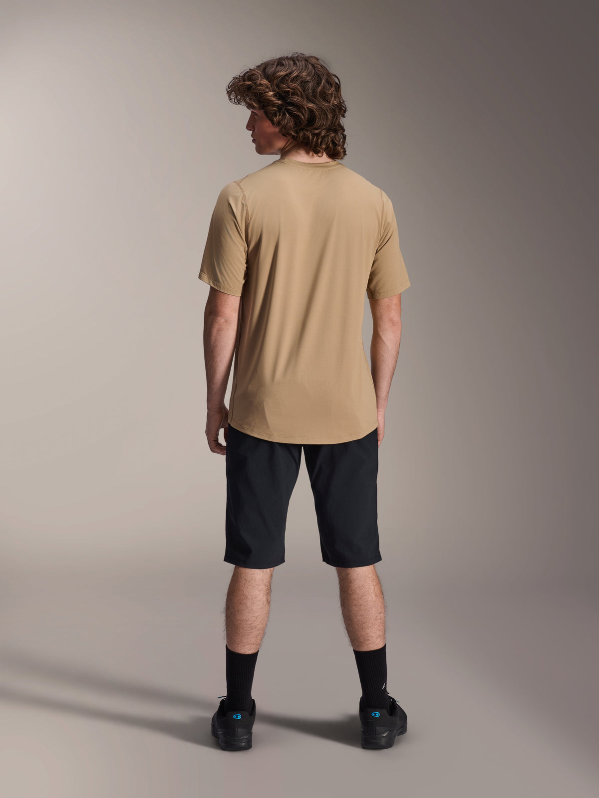 Ciclista masculino visto desde atrás con el jersey de manga corta Alpinestars A-Dura Astar en color marrón claro, combinado con pantalones cortos negros de mountain bike y calcetines negros, estilo casual para trail MTB.