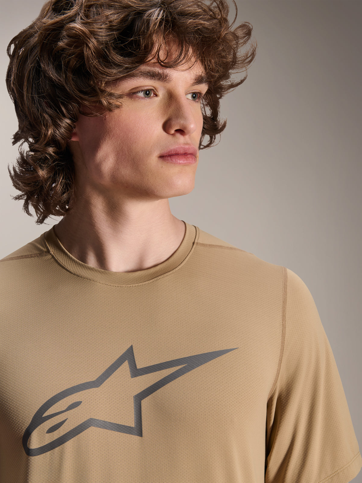 Modelo con el jersey Alpinestars A-Dura Astar de manga corta, jersey de MTB marrón claro con un logotipo de estrella gris en el pecho, tejido jacquard que absorbe la humedad, estilo de ciclismo casual