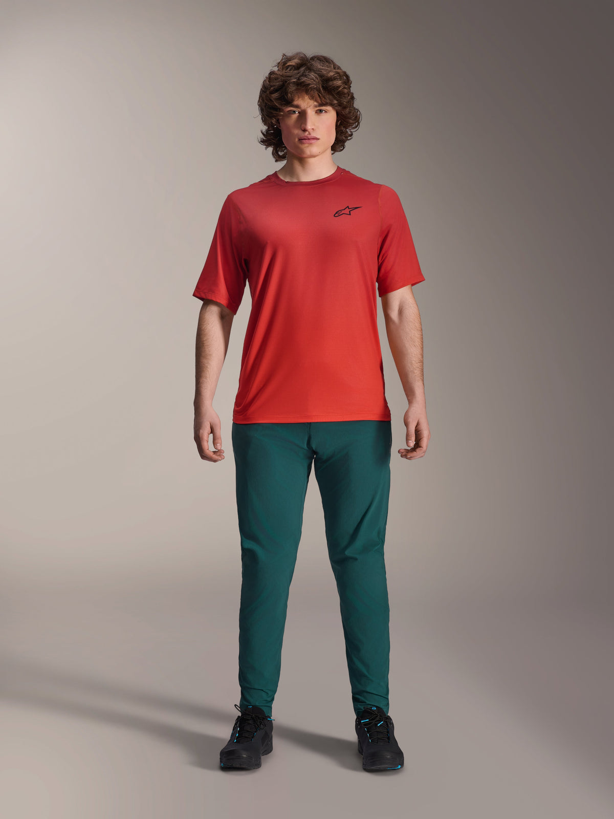 Alpinestars A-Dura Rise Jersey de manga corta, Spicy Orange, usado con pantalones verde oscuro y calzado negro, con un logotipo Astars negro en el pecho, ropa de ciclismo de montaña de alto rendimiento casual para conducción fuera de pista