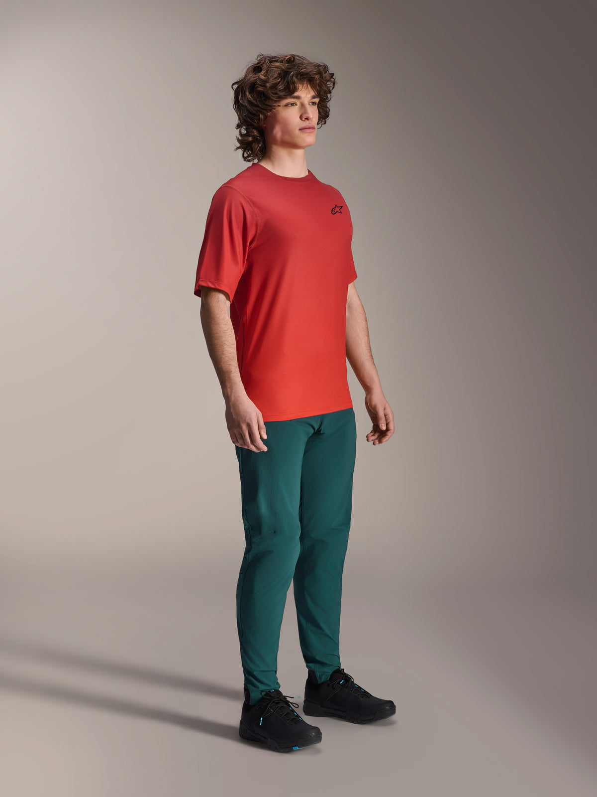 Model wearing Alpinestars A-Dura Pants in Dark Pine, paired with a red A-Dura Rise Short Sleeve Jersey and black MTB shoes, athletic mountain bike apparel in a studio setting