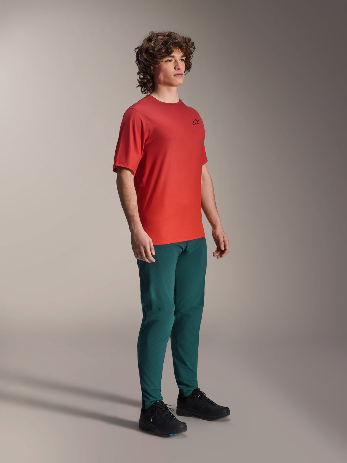 Camiseta Alpinestars A-Dura Rise de manga corta, color naranja Spicy Orange, con un pequeño logotipo negro en el pecho, combinada con pantalones verde azulado oscuro y zapatillas negras, ropa técnica de ciclismo de montaña ligera y transpirable