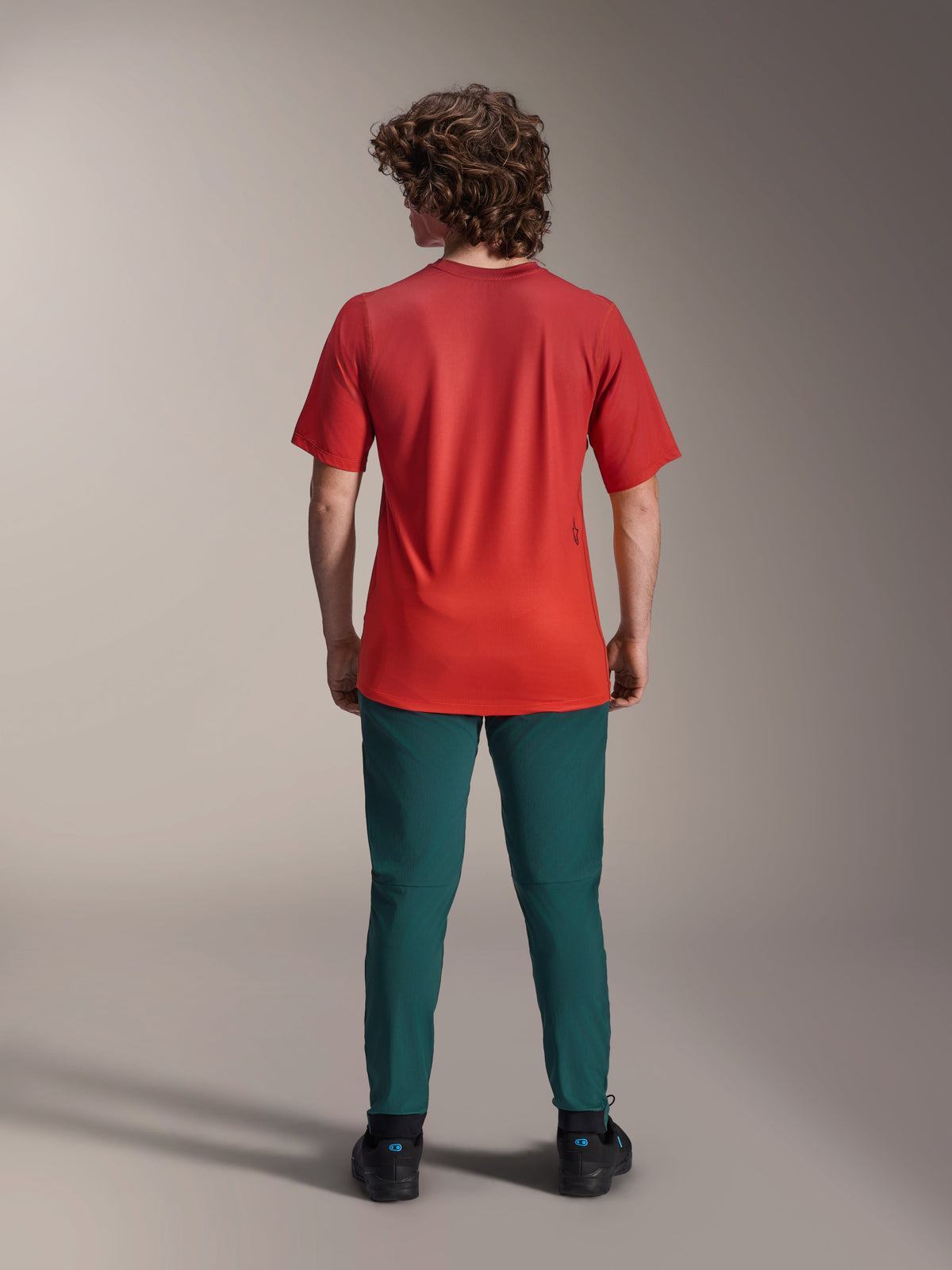 Alpinestars A-Dura Rise Jersey - Manga Corta, Spicy Orange, vista trasera de un modelo con pantalones verde azulado oscuro y calzado negro, con parte trasera más larga y un pequeño logotipo serigrafiado en la espalda, equipamiento ligero de alto rendimiento para MTB