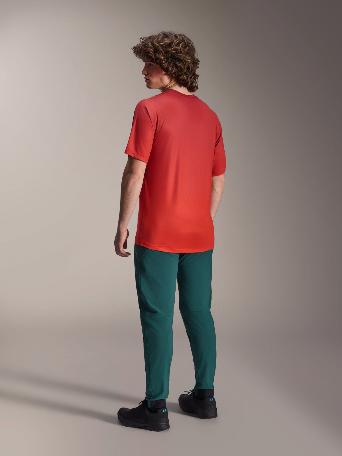 Alpinestars A-Dura Rise Jersey - Manga Corta en Spicy Orange, vista trasera del modelo con el jersey de ciclismo, pantalones de montaña verde oscuro y zapatillas negras, con una parte trasera más larga y ajuste de rendimiento para conducción todoterreno