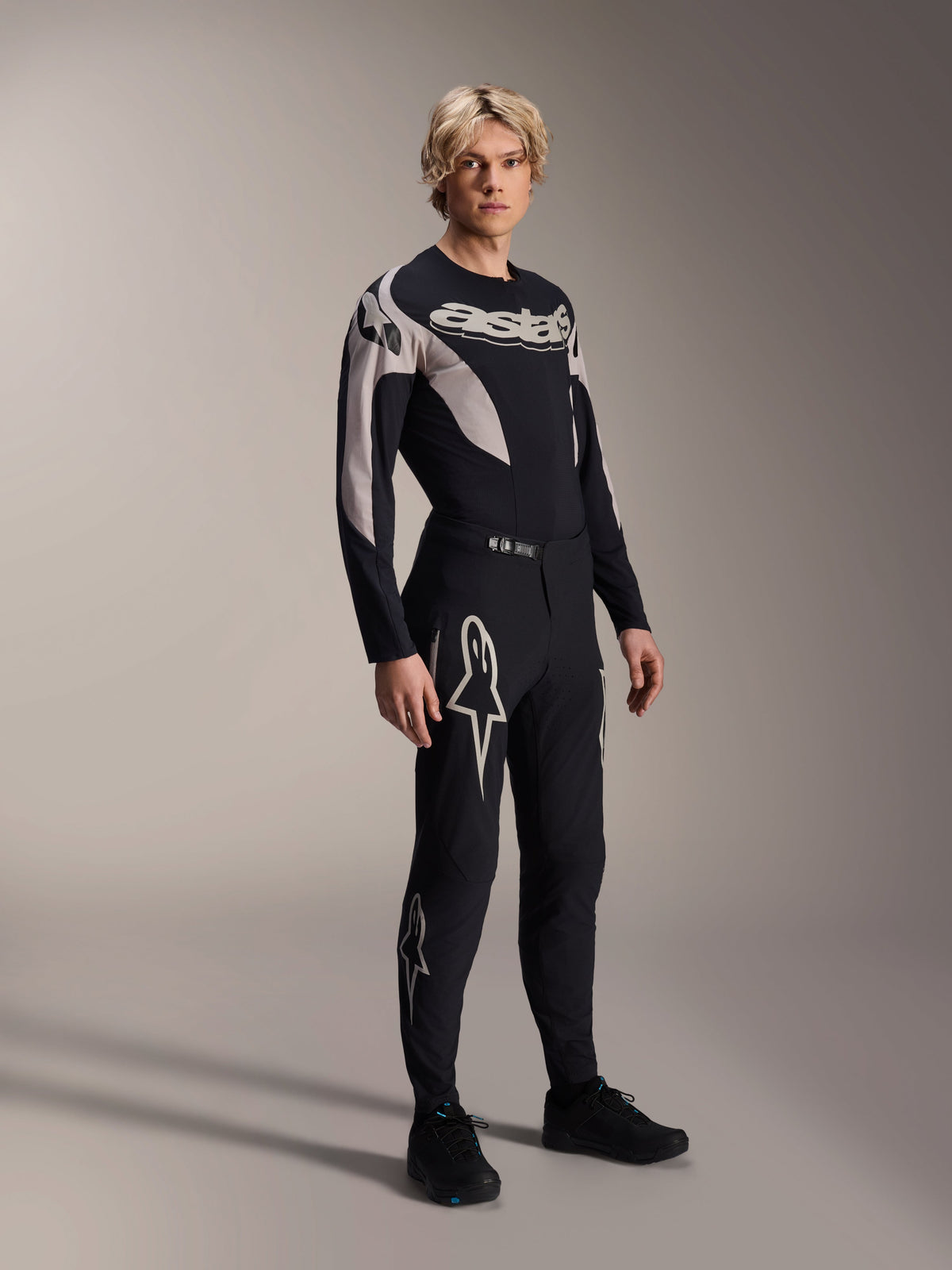 Modelo masculino vistiendo pantalones Alpinestars A-Supra Melt y jersey de manga larga a juego, negro con patrones curvos en gris claro, con destacados logotipos Astars blancos en las piernas y marca en el pecho, equipamiento profesional para ciclismo de montaña.