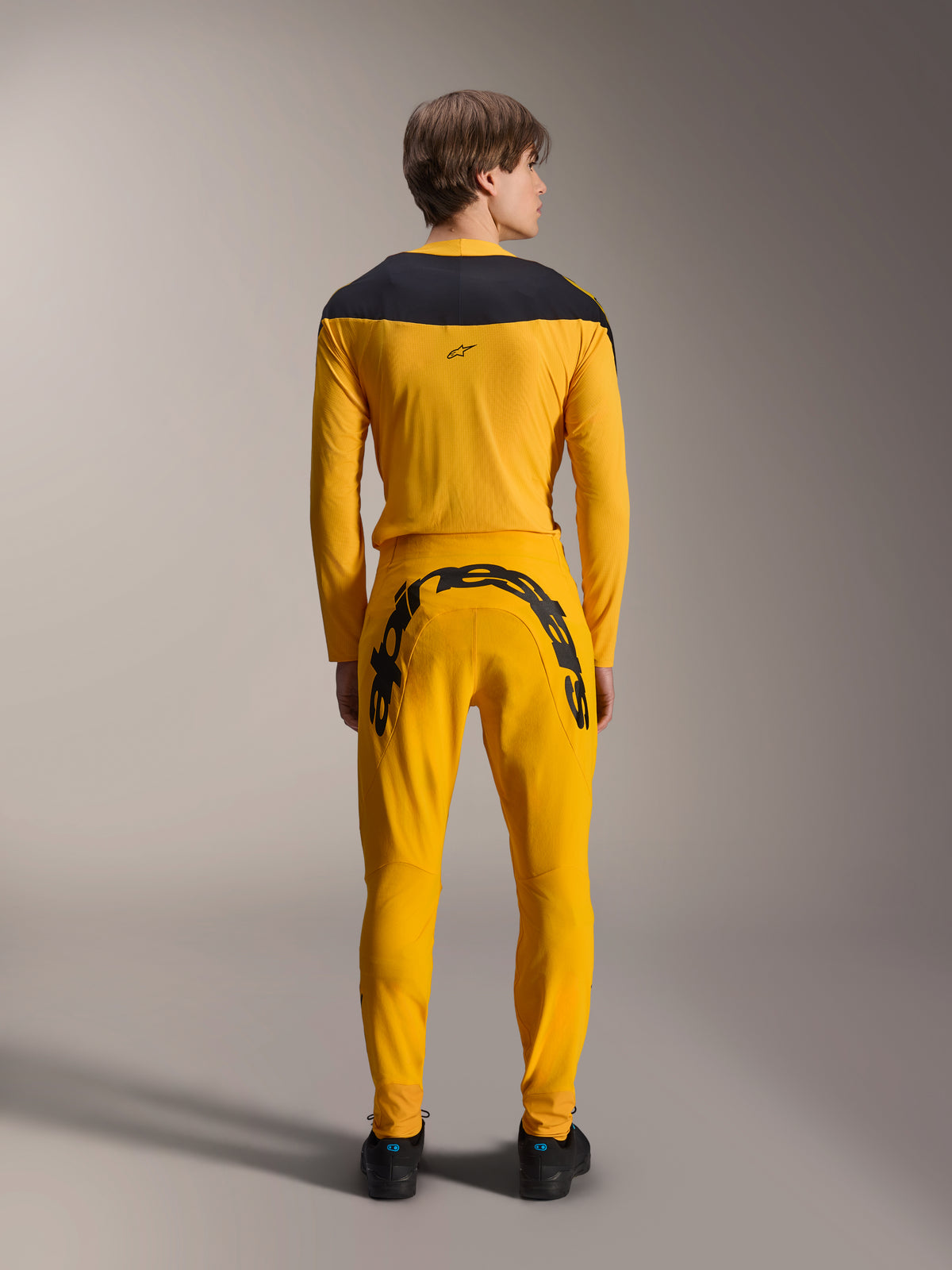 Rear view of a model wearing the Alpinestars Limited Edition A-Supra Melt Gold Gear-Set, featuring a dark yellow long-sleeve jersey with a black shoulder yoke and matching dark yellow pants with a large black Alpinestars wordmark across the back yoke, paired with black shoes.