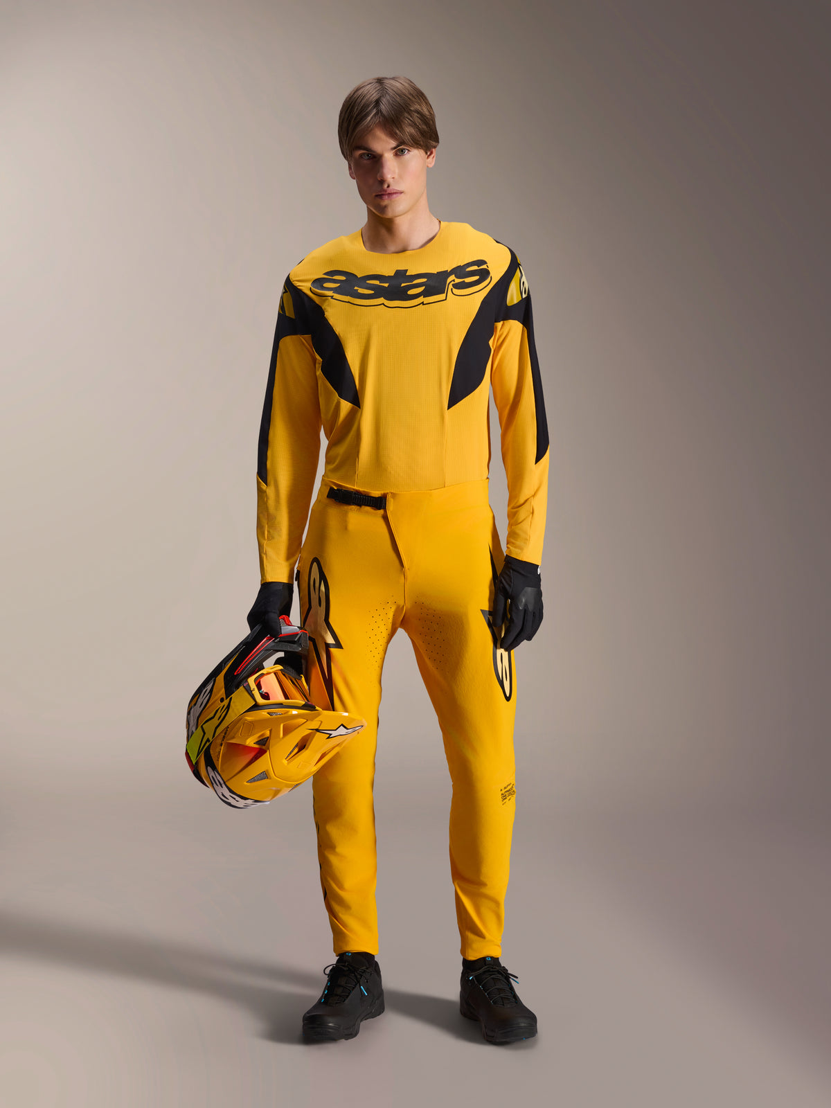 Model wearing Alpinestars Limited Edition A-Supra Melt Gold MTB Kit, yellow long-sleeve jersey with black 'astars' logo, matching yellow pants with black accents, holding a yellow and black racing helmet, black gloves and shoes, performance gear for mountain biking