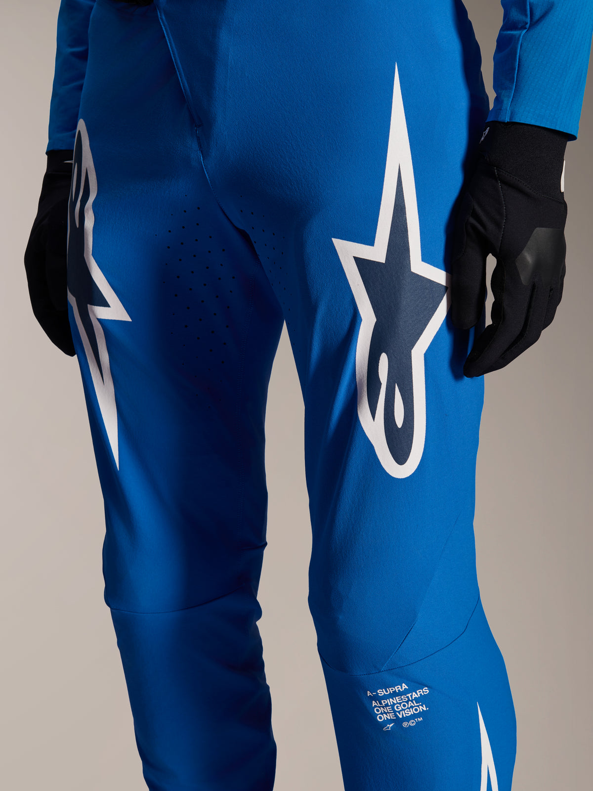 Alpinestars A-Supra Melt Pants, Blue cycling pants, featuring large white Astars logos on thighs, laser-cut perforations for airflow, and signature branding on the lower leg, worn with black gloves, performance MTB gear