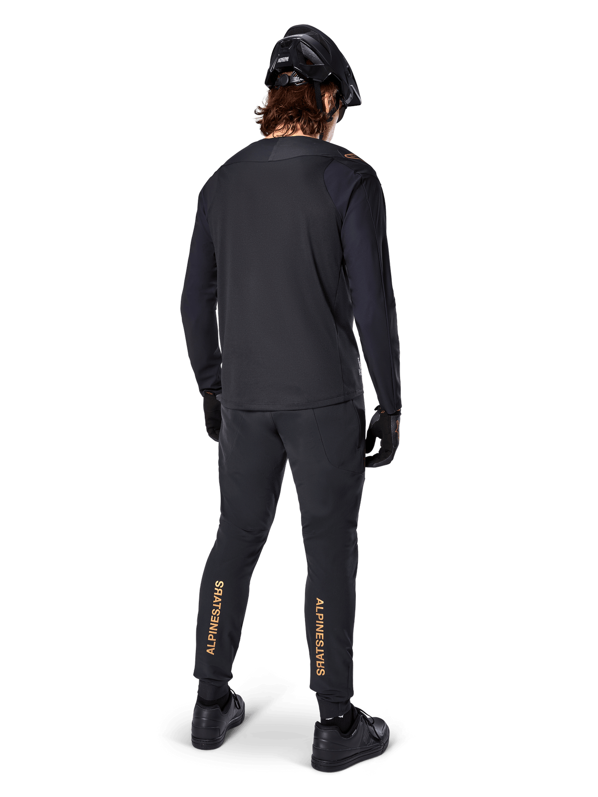 Rear view of a mountain bike rider wearing the Alpinestars A-Aria Polartec Switch Long Sleeve Jersey in Black, paired with black MTB pants featuring tan Alpinestars branding on the calves, black helmet and gloves, professional cycling gear for off-road trails.