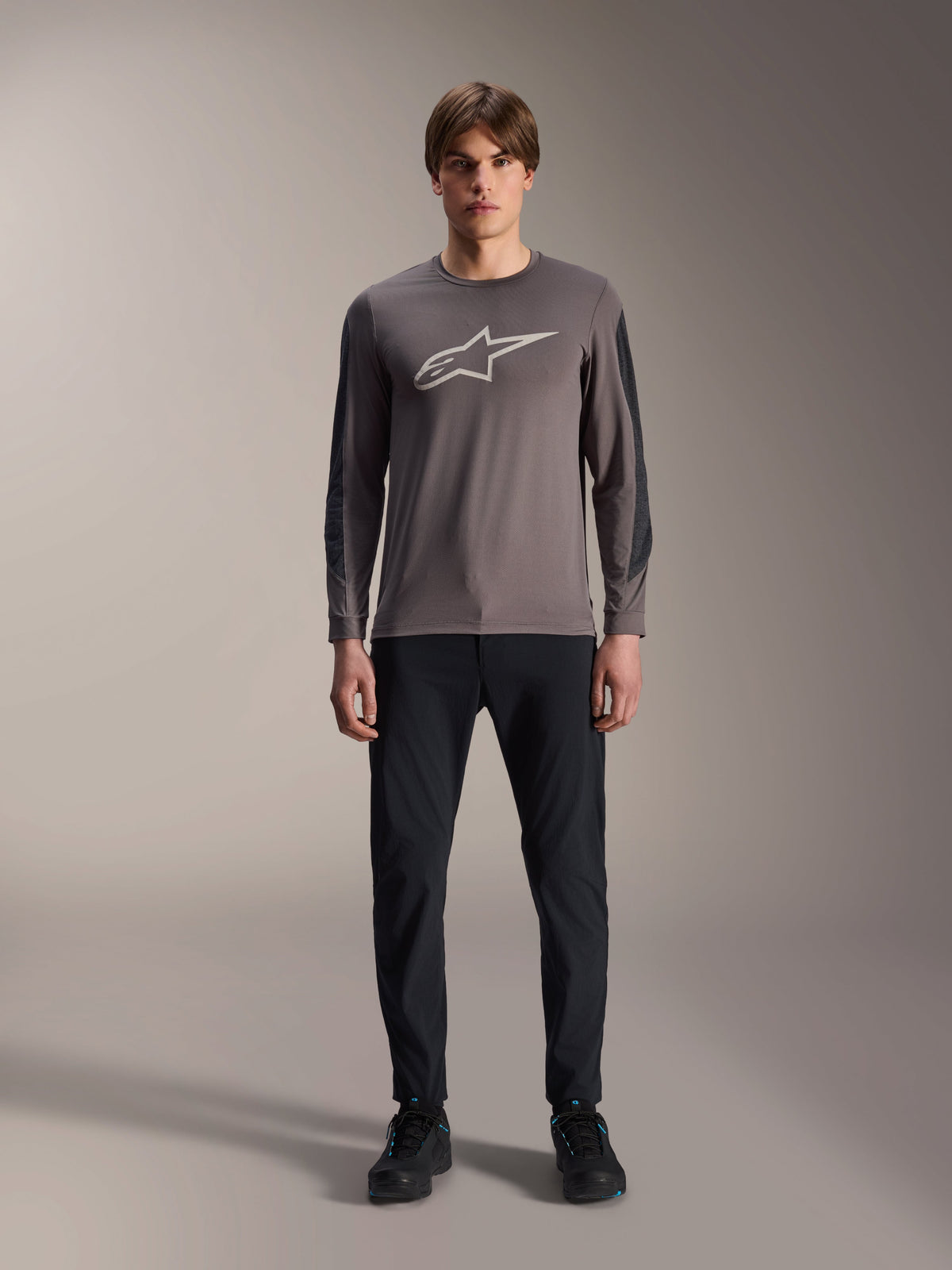 Alpinestars A-Dura Insert Astar Jersey, Long Sleeve MTB Jersey, Dark Gray, featuring a prominent light gray Astar logo on the chest and darker textured sleeve inserts, worn by a model with black pants and shoes, performance gear for mountain biking