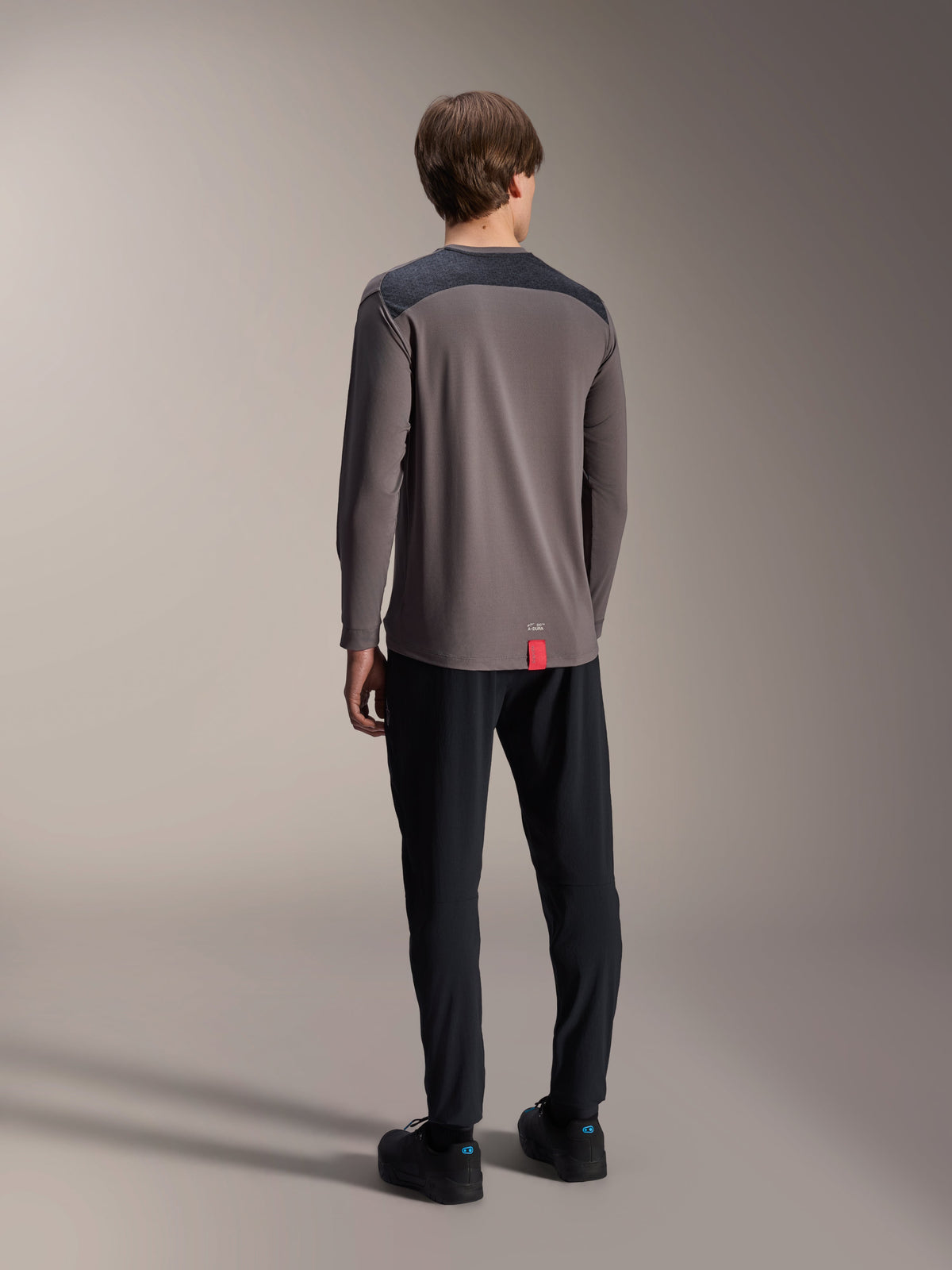 Alpinestars A-Dura Insert Astar Long Sleeve Jersey, Dark Gray, rear view on model, featuring charcoal reinforced shoulder panels and a small red hem tag, paired with black pants and shoes, technical MTB apparel