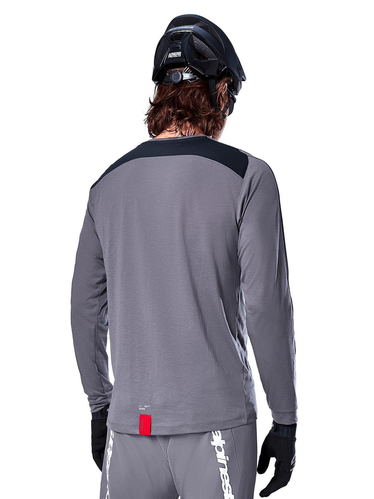 Alpinestars A-Dura Dri Astar Long Sleeve Jersey, jersey de MTB gris oscuro visto desde atrás, con un panel negro en los hombros y una pequeña etiqueta roja de la marca, usado con un casco de bicicleta de montaña y guantes negros.