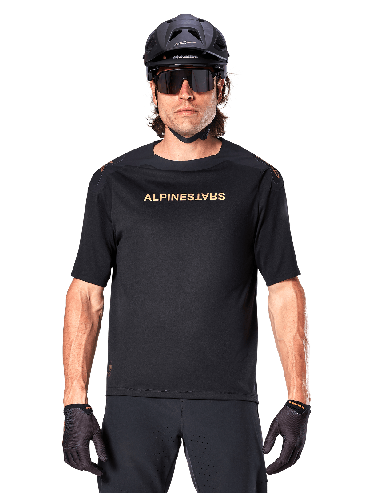 Mountain biker wearing Alpinestars A-Aria Polartec Switch Short Sleeve Jersey in Black, featuring gold-toned chest branding, paired with a black MTB helmet, dark performance sunglasses, and matching gloves, off-road cycling gear.