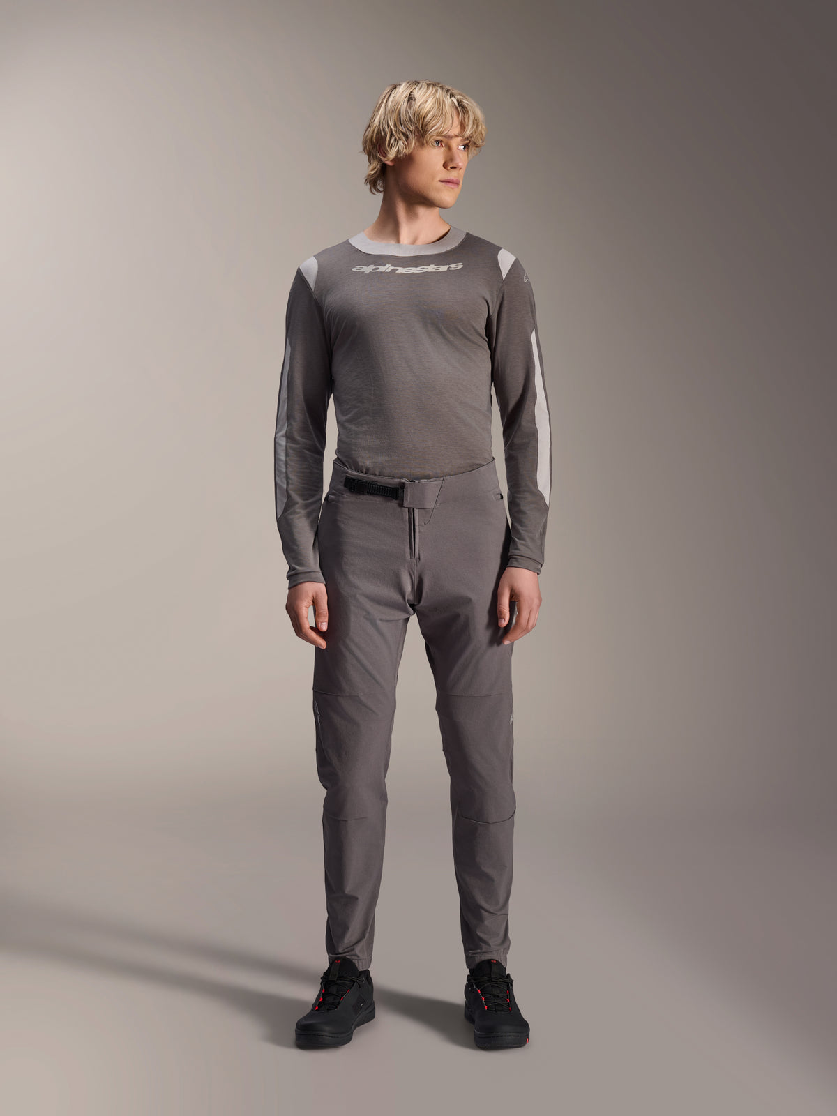 Pantalones Alpinestars A-Dura Elite, pantalones de ciclismo de montaña gris oscuro, vista de cuerpo completo, usados con un jersey de rendimiento de manga larga gris a juego con el logo de Alpinestars, zapatillas negras con detalles rojos, conjunto de ciclismo deportivo