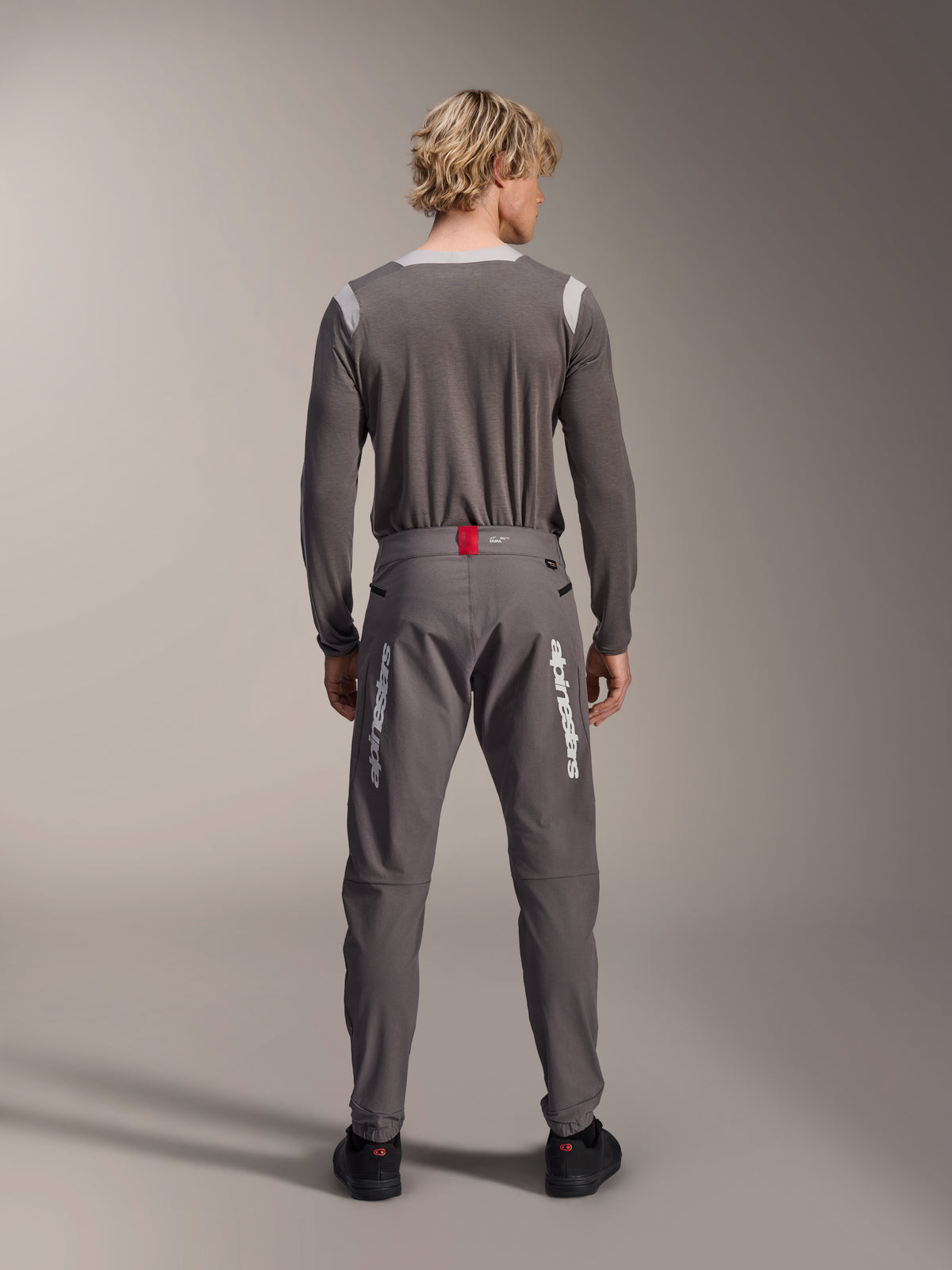Modelo de espaldas vistiendo pantalones Alpinestars A-Dura Elite en gris oscuro y un jersey de manga larga gris a juego, con logotipos blancos de Alpinestars en las piernas, un detalle rojo en la cintura y zapatillas negras, equipamiento profesional para ciclismo MTB.