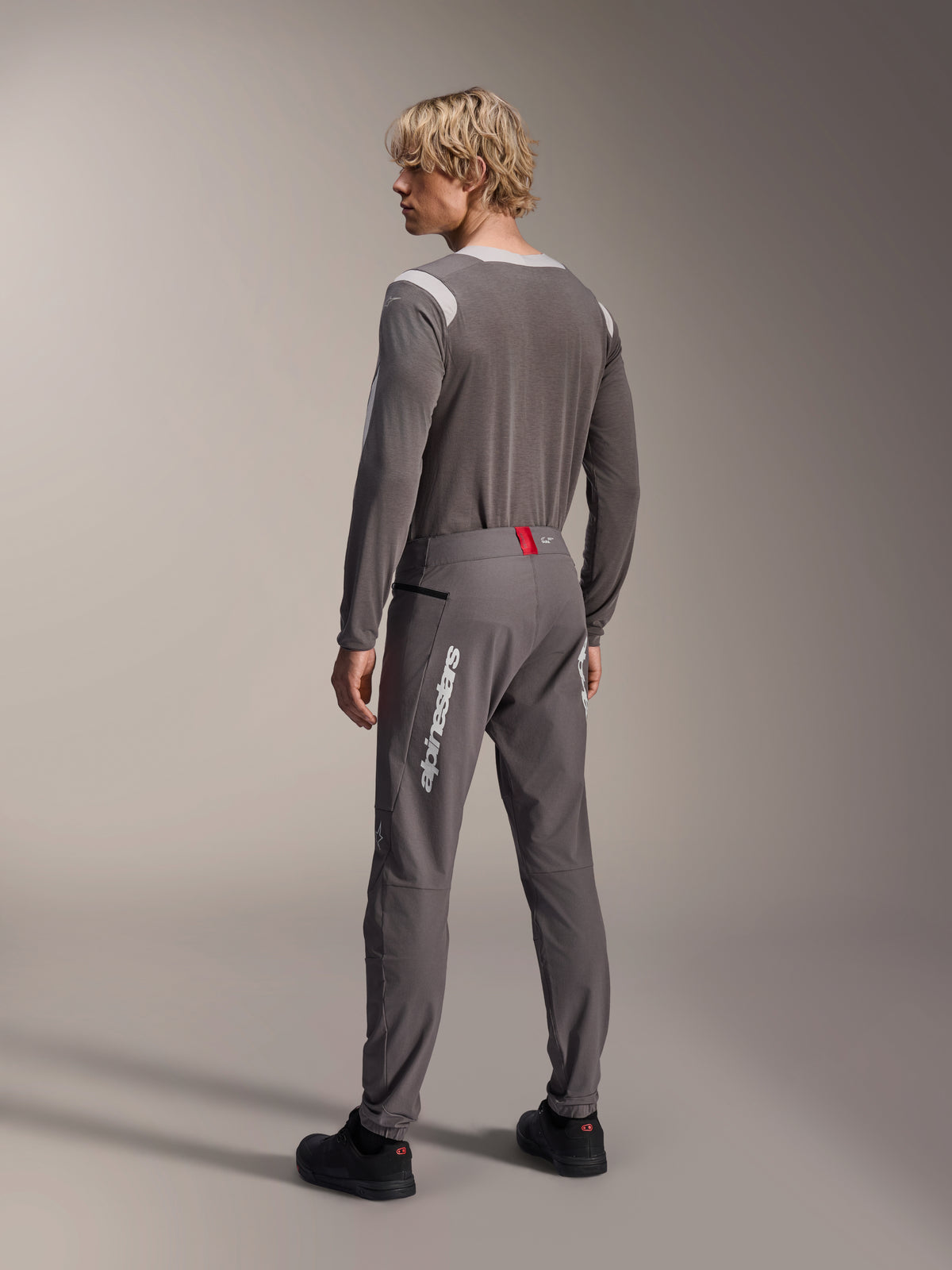 Modelo desde una vista posterior con equipamiento MTB de Alpinestars, jersey de manga larga A-Dura Dri Wool gris, pantalones A-Dura Elite gris oscuro con marca vertical blanca y detalle rojo en la cintura, conjunto de ciclismo coordinado para mountain bike