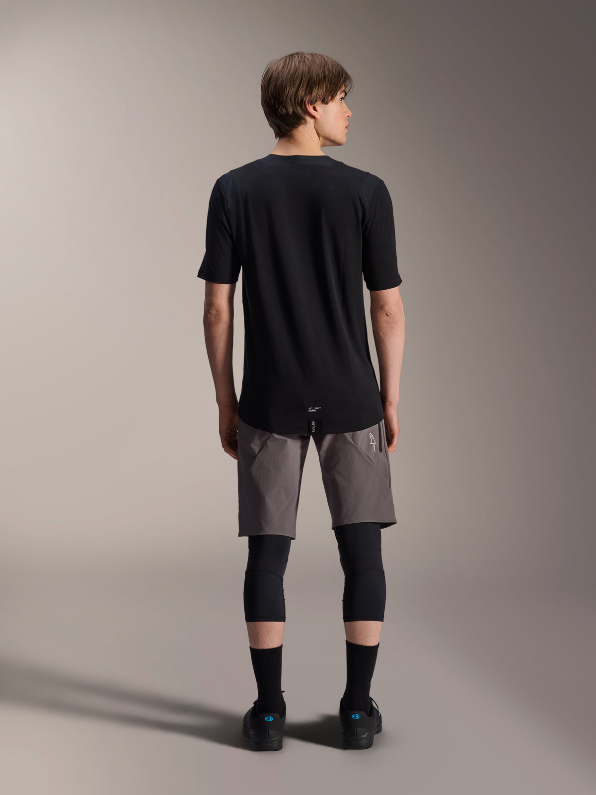 Alpinestars A-Dura Elite Shorts, Dark Gray mountain bike shorts, back view, worn with a black short-sleeve jersey, knee pads, and black socks, technical cycling gear for off-road riding