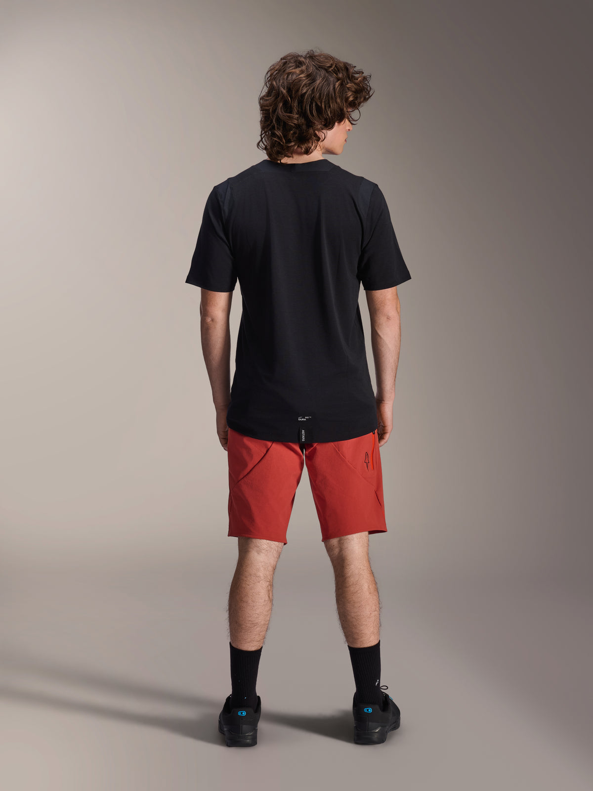 Modelo masculino vistiendo pantalones cortos Alpinestars A-Dura Elite en color Dark Orange y un jersey de manga corta A-Dura Dri Wool negro, visto desde atrás, combinado con calcetines negros y zapatillas de ciclismo, ropa profesional para bicicleta de montaña