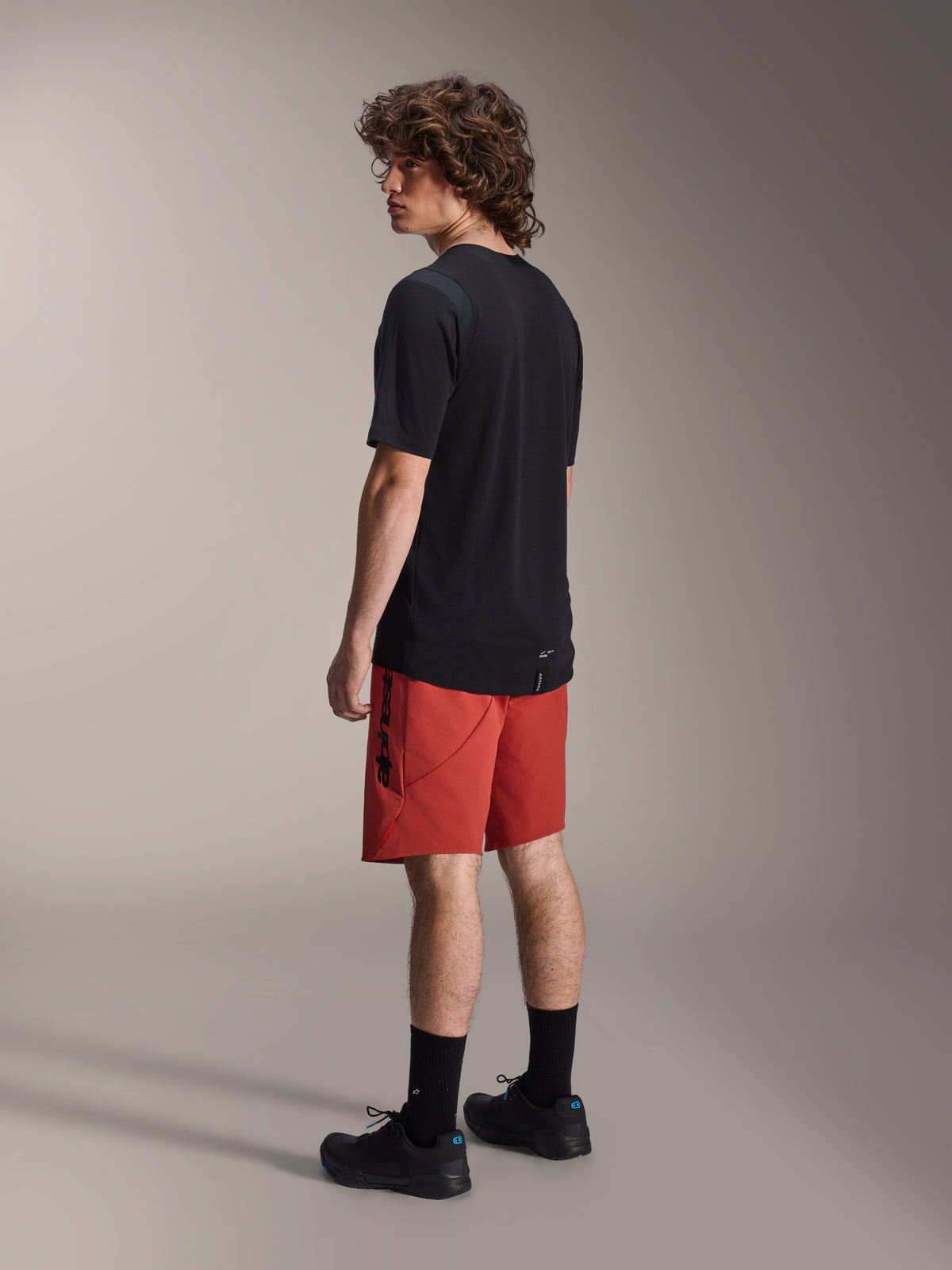 Shorts Alpinestars A-Dura Elite, pantalones cortos de mountain bike, naranja oscuro, usados con un jersey negro de manga corta y zapatillas de mountain bike negras, vista lateral trasera del modelo destacando el ajuste de alto rendimiento y la marca