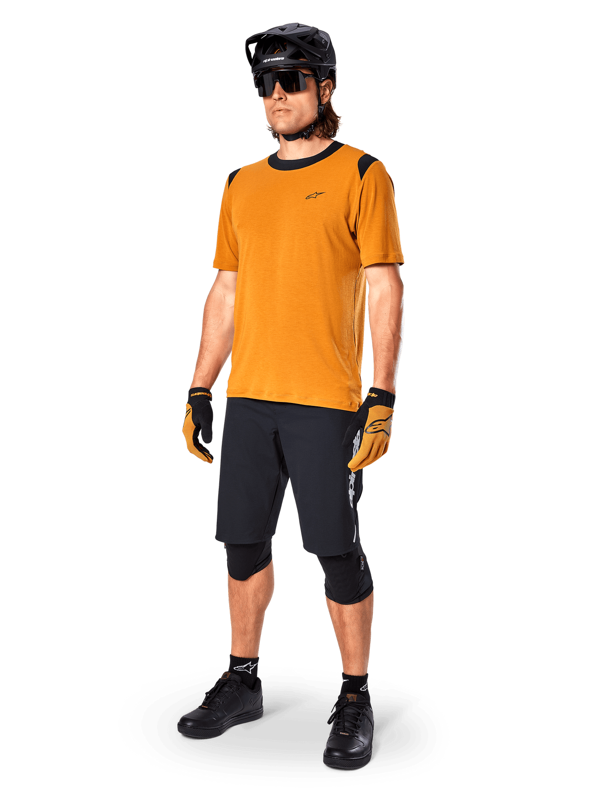Modelo masculino vistiendo el Jersey Alpinestars A-Dura Dri Wool - Manga Corta en color Dorado Oscuro, jersey de ciclismo de montaña con detalles negros en hombros y cuello, combinado con pantalones cortos de MTB negros, casco negro y gafas de sol, equipamiento técnico de ciclismo todoterreno