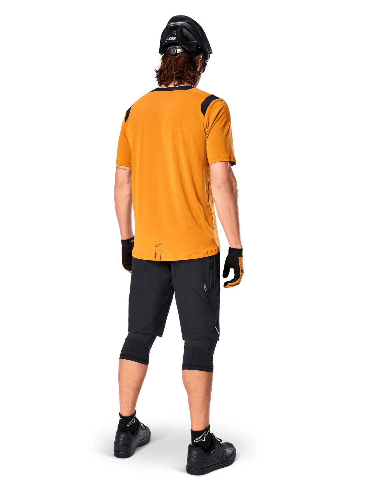 Alpinestars A-Dura Dri Wool Short Sleeve Jersey, jersey de ciclismo, oro oscuro, vista trasera de ciclista de montaña con jersey amarillo mostaza y acentos de malla negra en los hombros, combinado con pantalones cortos negros, rodilleras, casco negro y guantes, equipamiento técnico de ciclismo todoterreno