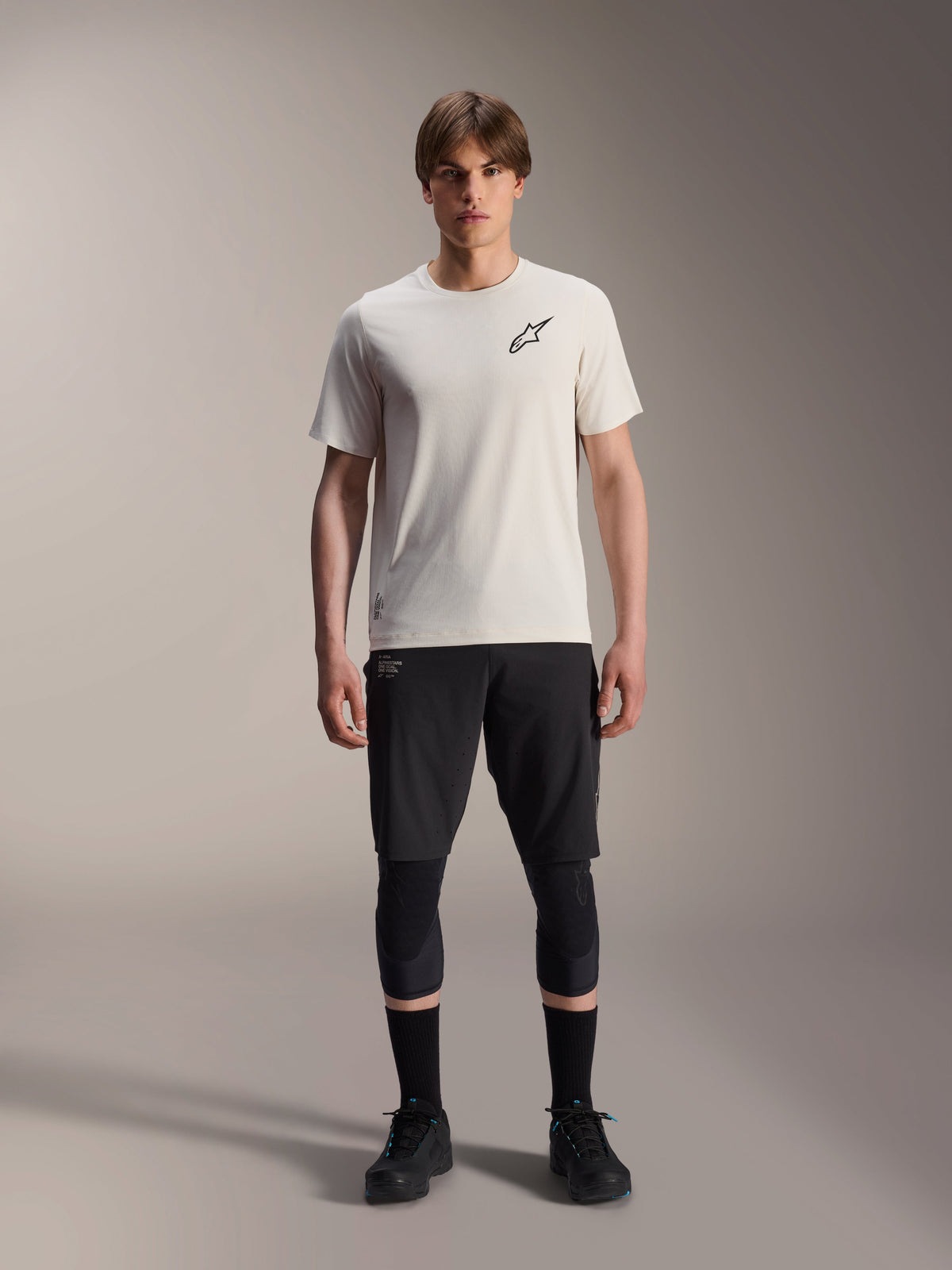 Male model wearing the Alpinestars A-Aria Astar Short Sleeve Jersey in Ecru, paired with black MTB shorts, knee guards, and black cycling shoes, featuring a minimalist black star logo on the chest, designed for off-road trail riding.