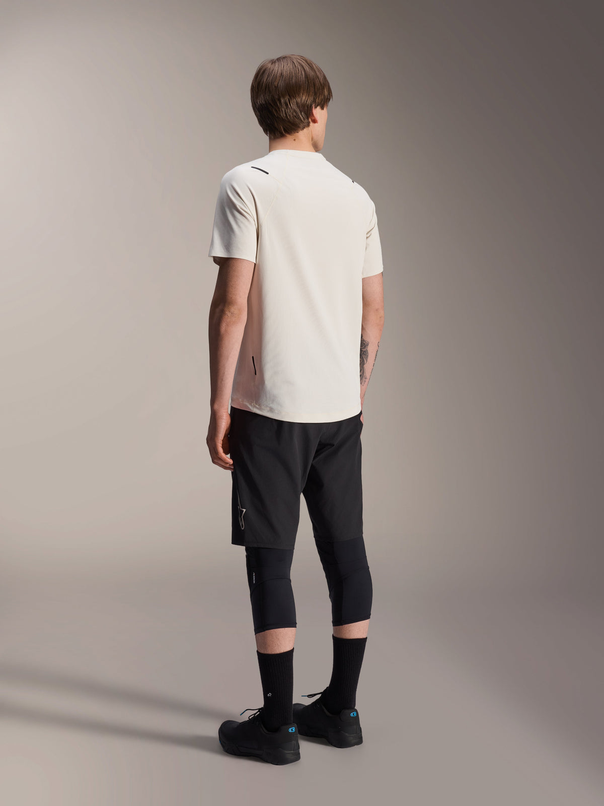 Alpinestars A-Aria Astar Jersey, Short Sleeve MTB Jersey, Ecru, back view showing the performance fit and longer drop tail, styled with black mountain bike shorts, knee protectors, and black riding shoes for off-road trail sessions