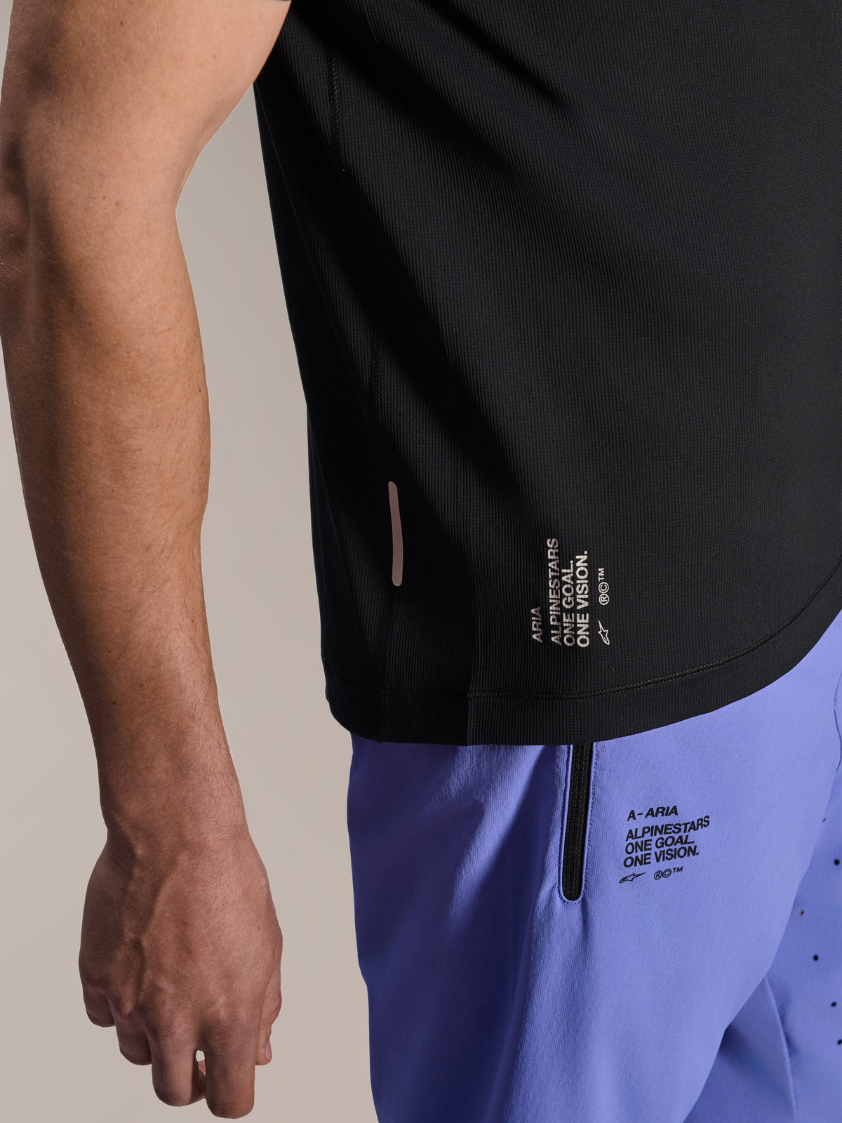 Jersey de manga corta Alpinestars A-Aria Switch, jersey de MTB negro, primer plano del dobladillo lateral con la marca blanca 'ONE GOAL. ONE VISION.', usado con pantalones cortos de ciclismo de color morado claro con logotipos a juego