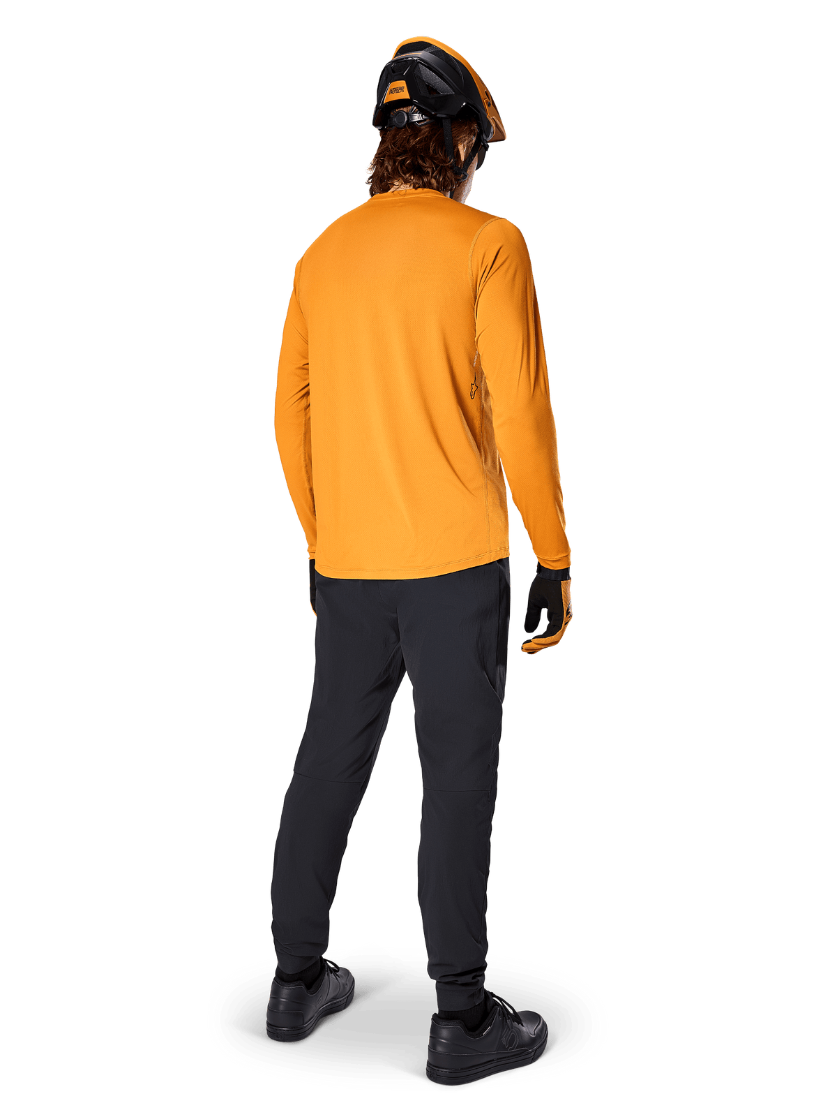 Ciclista de montaña visto desde atrás con el jersey de manga larga Alpinestars A-Dura Rocker en color Dark Gold, pantalones negros, casco naranja y negro, y zapatillas negras, equipamiento de MTB de alto rendimiento para recorrer senderos